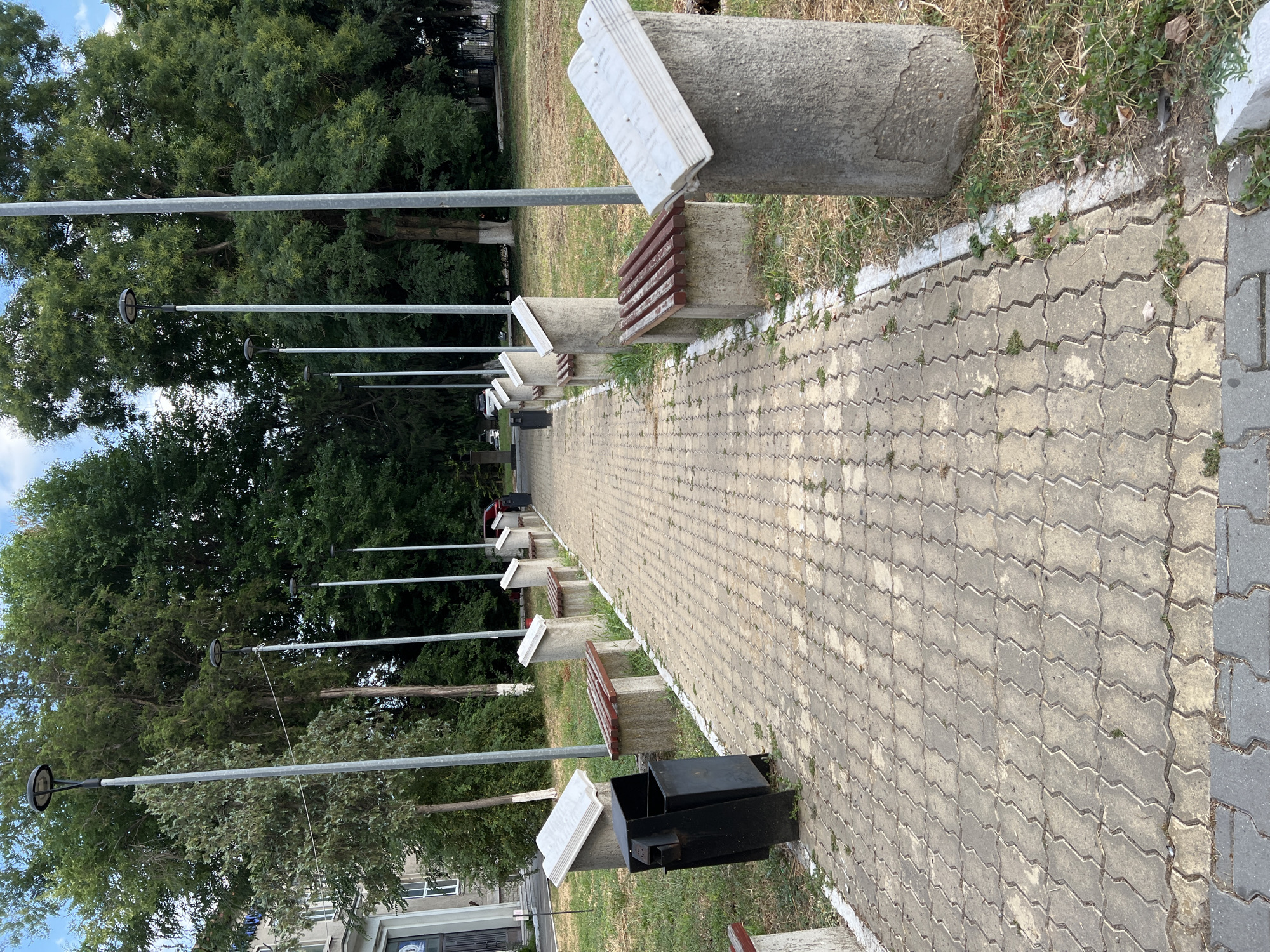 The image shows a paved walkway lined with benches and concrete structures that resemble open books. Streetlights are visible along the walkway, and trees and vegetation are present in the background. The scene appears to be an outdoor public space, possibly a park or a memorial area, in the city of Constanța, Romania. A black trash can is located near the beginning of the walkway. The overall condition of the area suggests it is well-maintained, though some grass is visible growing through the paving stones and the surrounding soil.