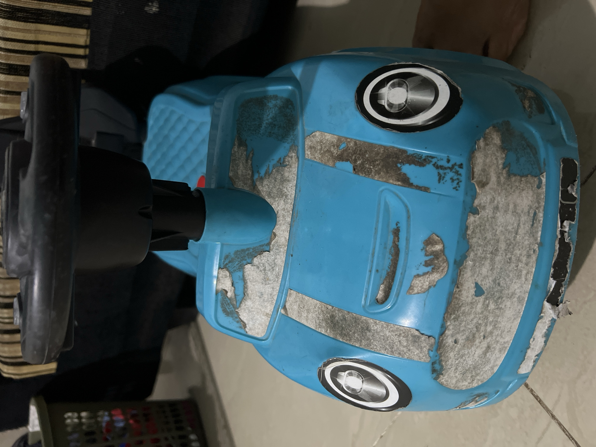 A well-worn, blue plastic ride-on toy car sits on a tiled floor. The car has a black steering wheel and a silver stripe design on its hood, which is peeling in several places. The headlights are represented by circular stickers with a reflective interior. The toy is positioned in what appears to be a residential setting in Klapanunggal, Indonesia, with a basket and furniture visible in the background. The lighting suggests it is indoors, likely in a home.