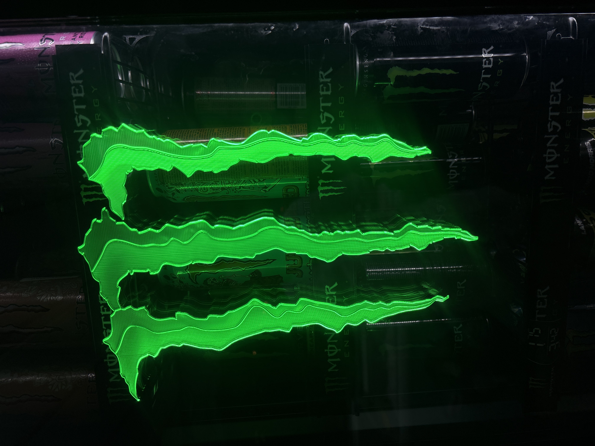 The image displays a shelf filled with Monster Energy drinks. The green Monster logo is prominently lit, creating a neon-like effect. The cans are various flavors of Monster, evidenced by the partial views of labels such as "Juice" and "Ultra". The image was taken in Plovdiv, Bulgaria.
