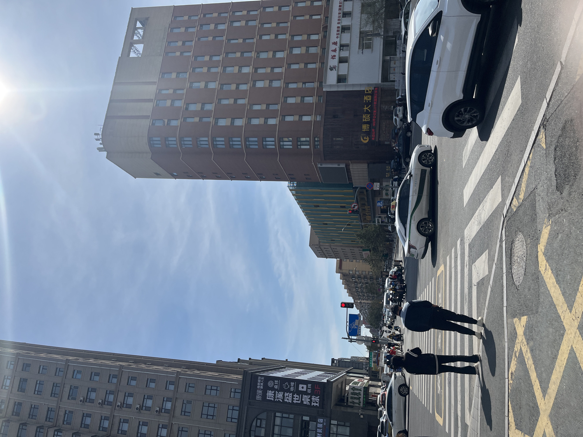 The image depicts a street scene in Changchun City, China, featuring a tall, brown building dominating the background. The sky is partly cloudy with a bright sun shining. In the foreground, two pedestrians are walking across a crosswalk. Cars are parked along the street and are stopped at a red light. A large sign with Chinese characters and English letters "3F" is visible on one of the buildings. The street is marked with yellow lines and the crosswalk has white stripes. A restaurant, named "Bo Shuo Restaurant" is visible in the scene.