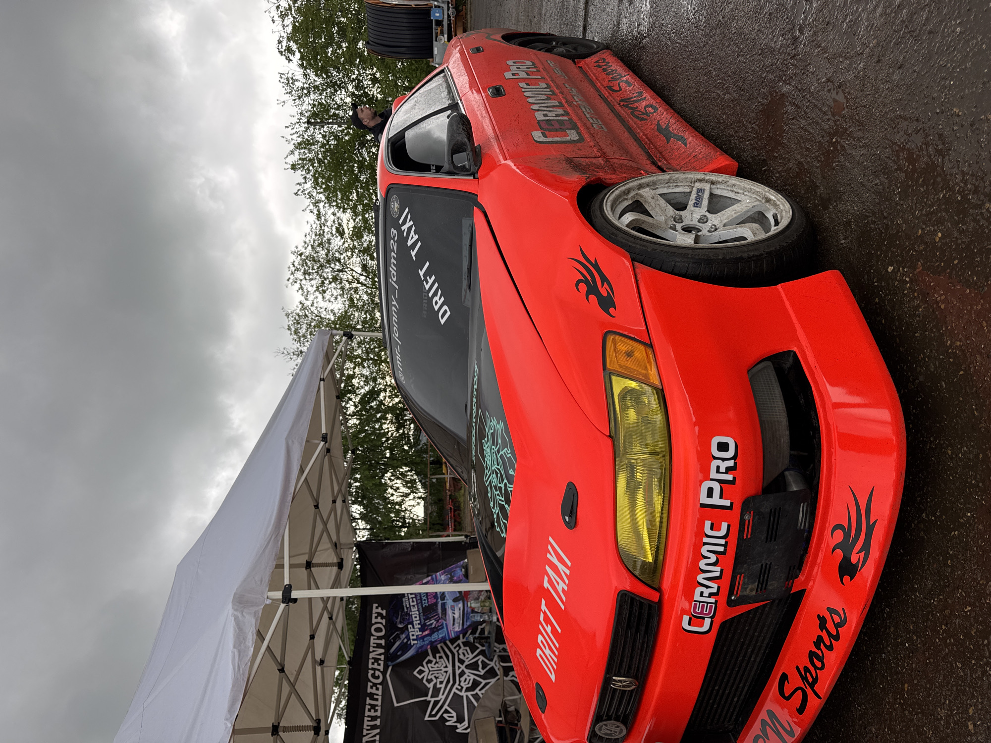 A bright orange drift car is angled in the image, with a white tent partially visible to the left. The car features decals like "DRIFT TAXI" and "CERAMIC PRO," along with tribal flame designs. It's parked on wet pavement, suggesting a recent rain. The sky is overcast with grey clouds. A person is visible inside the car. A banner that reads "NTELEGENTOFF" is partially shown. A tire is near the back of the car on the road. The photo was taken in Pavlovskaya, Russia.