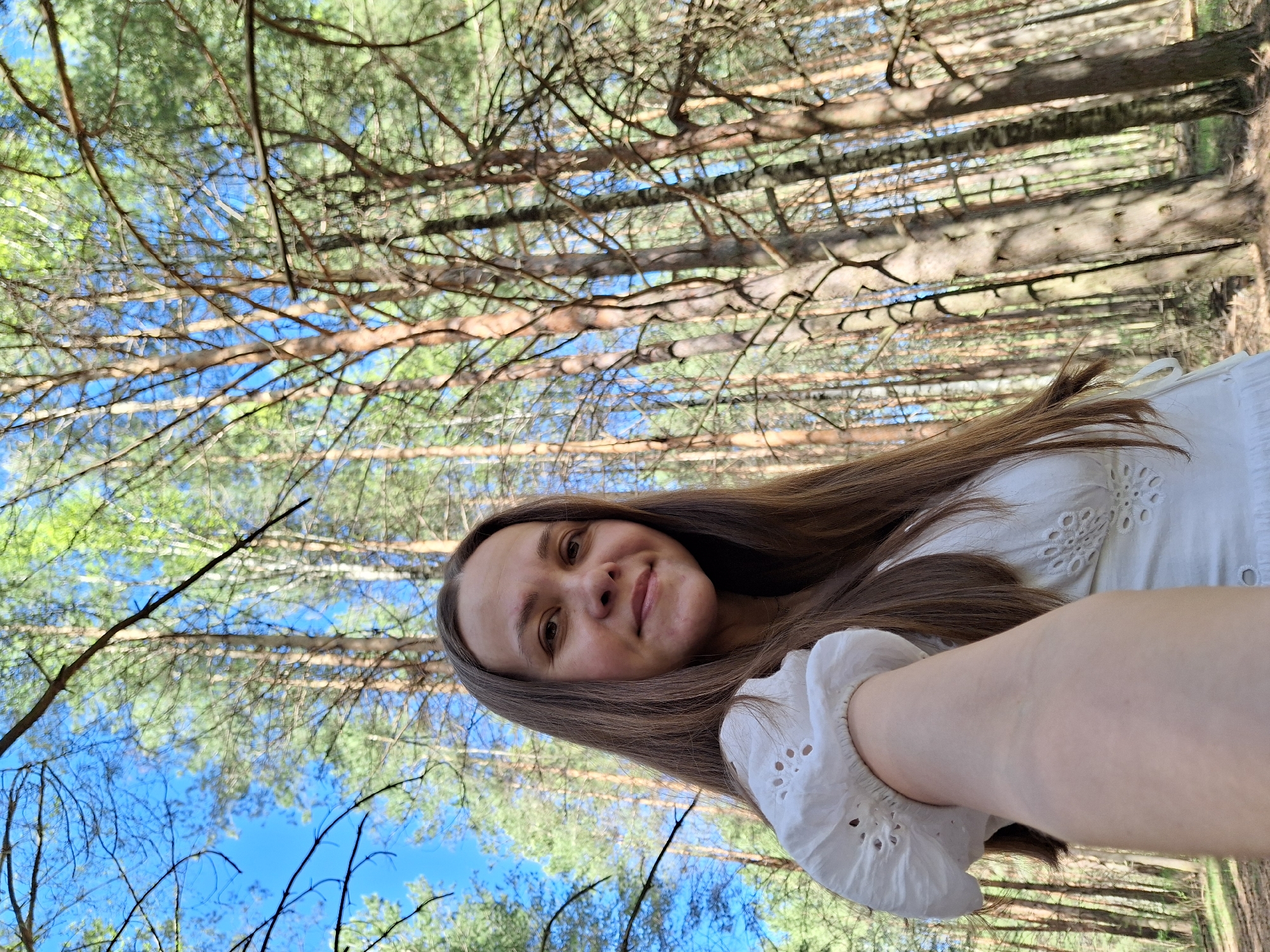 A young woman with long brown hair is taking a selfie in a forest. She is wearing a white, eyelet-detailed dress. The background reveals tall trees with branches against a blue sky. Based on the prompt information, this is likely a wooded area near Chaikovsky, Russia.