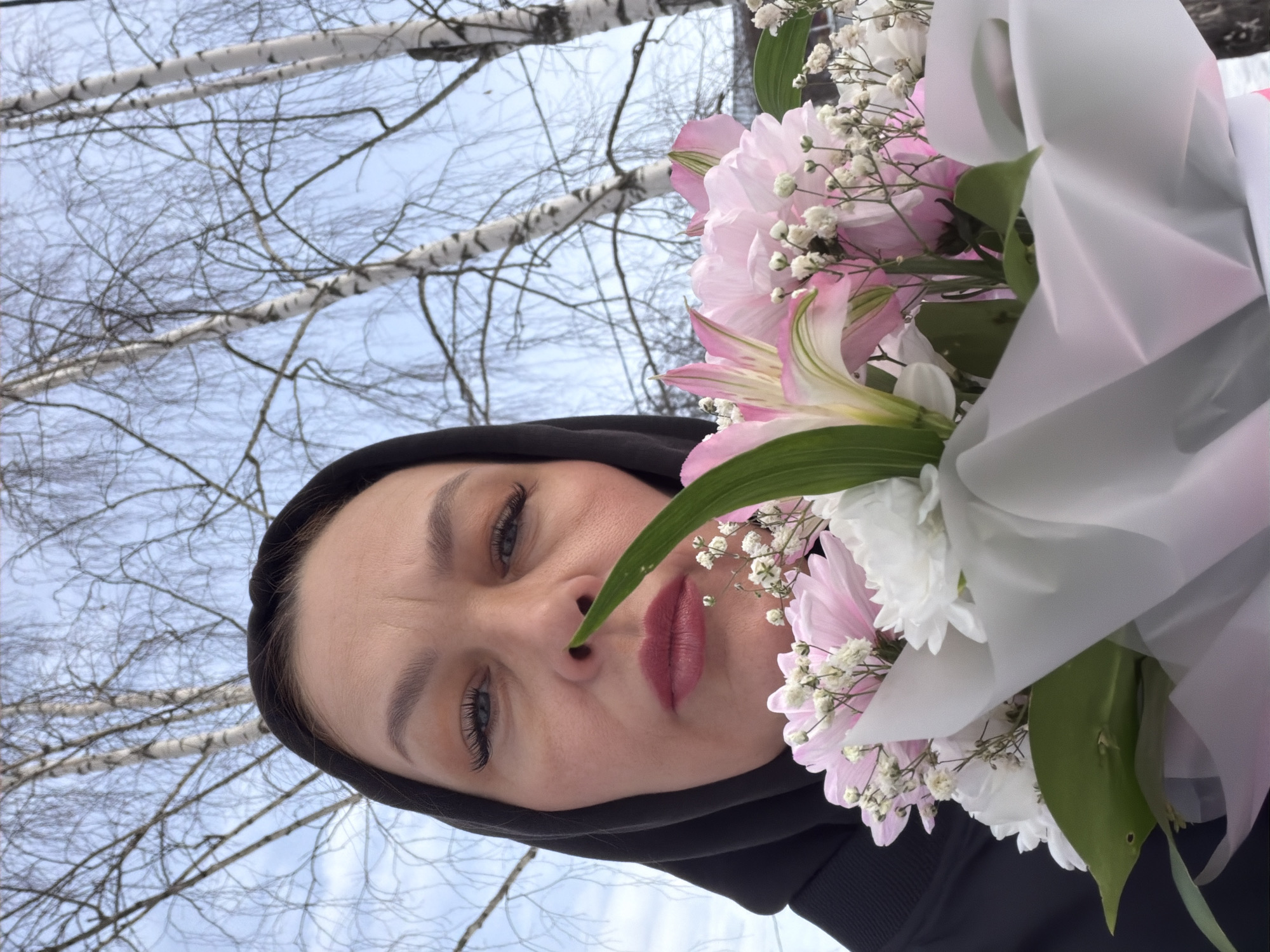 A close-up selfie photograph depicts an adult female holding a bouquet of flowers outdoors in Chaikovsky, Russia. The woman, positioned in the lower central frame, wears a black hooded garment that covers her head. Her visible facial features include dark eyebrows, prominent eyelashes, and pink-toned lipstick. She looks directly at the camera. A bouquet of flowers is held in the foreground, partially obscuring her lower face. The bouquet comprises pink alstroemeria, large pink chrysanthemums, smaller white chrysanthemums, and white baby's breath, wrapped in white and light pink material. A single green leaf from the bouquet extends upwards, positioned in front of the woman's nose. The background features several leafless birch trees with white bark and slender branches against a pale blue, overcast sky, indicating a cold weather season.