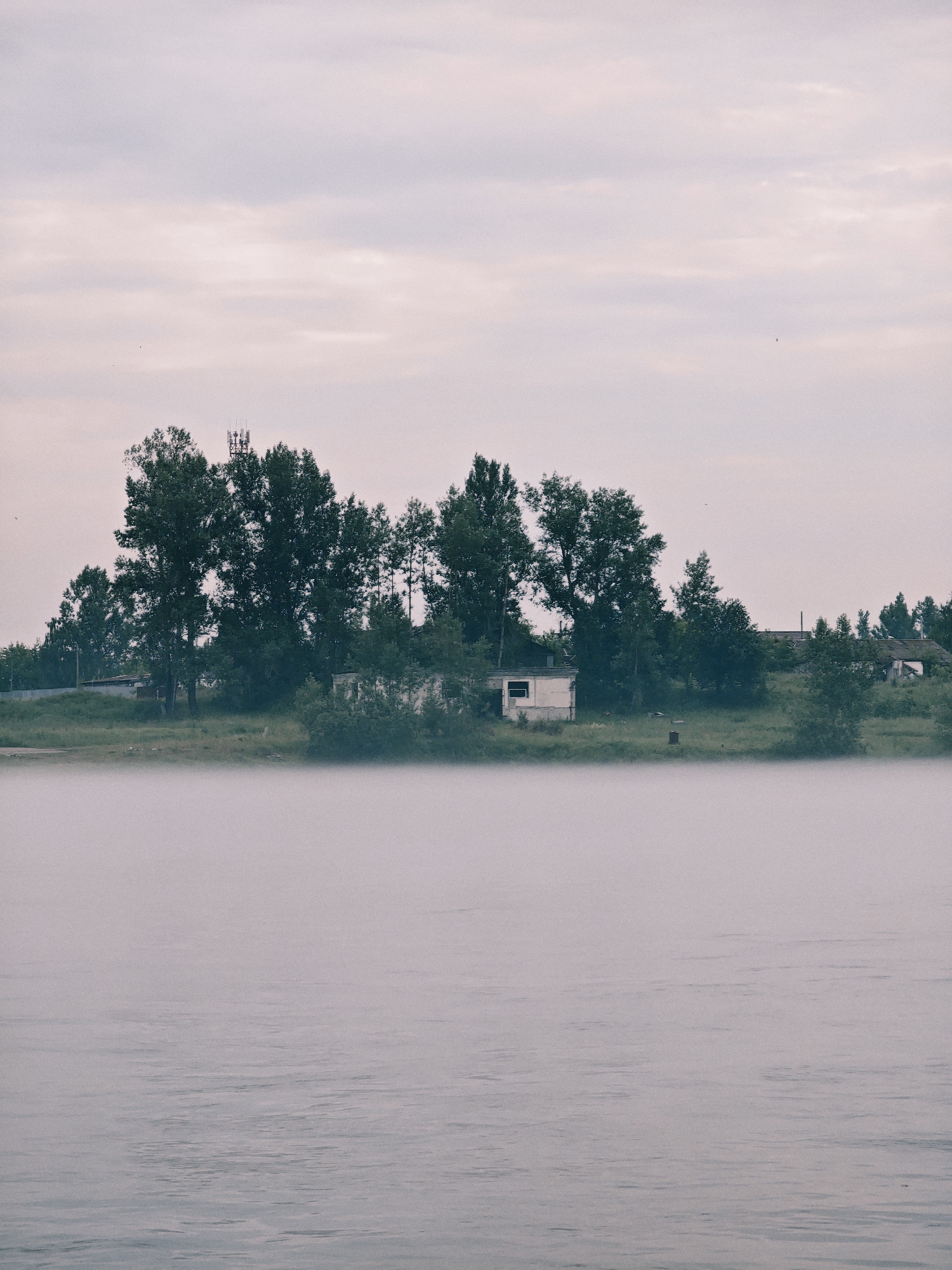 A wide shot captures a foggy river in Zheleznogorsk, Russia, under a pale sky. The foreground is dominated by the misty river, its surface disturbed by gentle ripples. In the midground, a strip of land separates the river from a line of trees. Among the trees, several abandoned buildings are visible, including a small, dilapidated structure with a single window on the left and other longer, low-rise buildings further back, one with a broken roof. A tall radio tower stands behind the trees on the left. The scene suggests a tranquil, somewhat desolate, natural environment at the edge of a settlement.