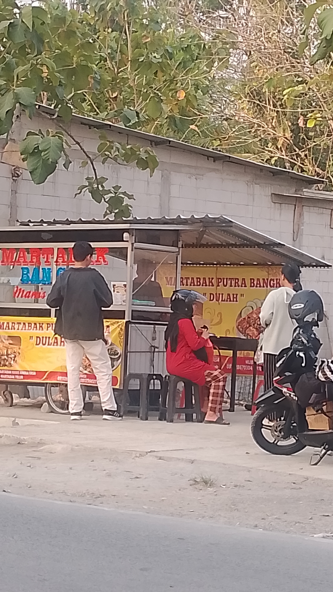 The image depicts an outdoor food stall in Karanglangu, Indonesia. A vendor, a man wearing a dark jacket and light pants, is standing at the counter of the stall, which is adorned with signs that read "MARTABAK PUTRA BANG" and "DULAH". To his right, a woman in a red shirt and plaid pants sits on a stool, wearing a helmet. Behind her, another person in a white shirt and striped pants is standing. A motorcycle is parked to the right of the scene. The stall is set up in front of a concrete building. The ground is a dirt and gravel surface, and vegetation is visible in the background. The lighting suggests it is late afternoon or early evening.