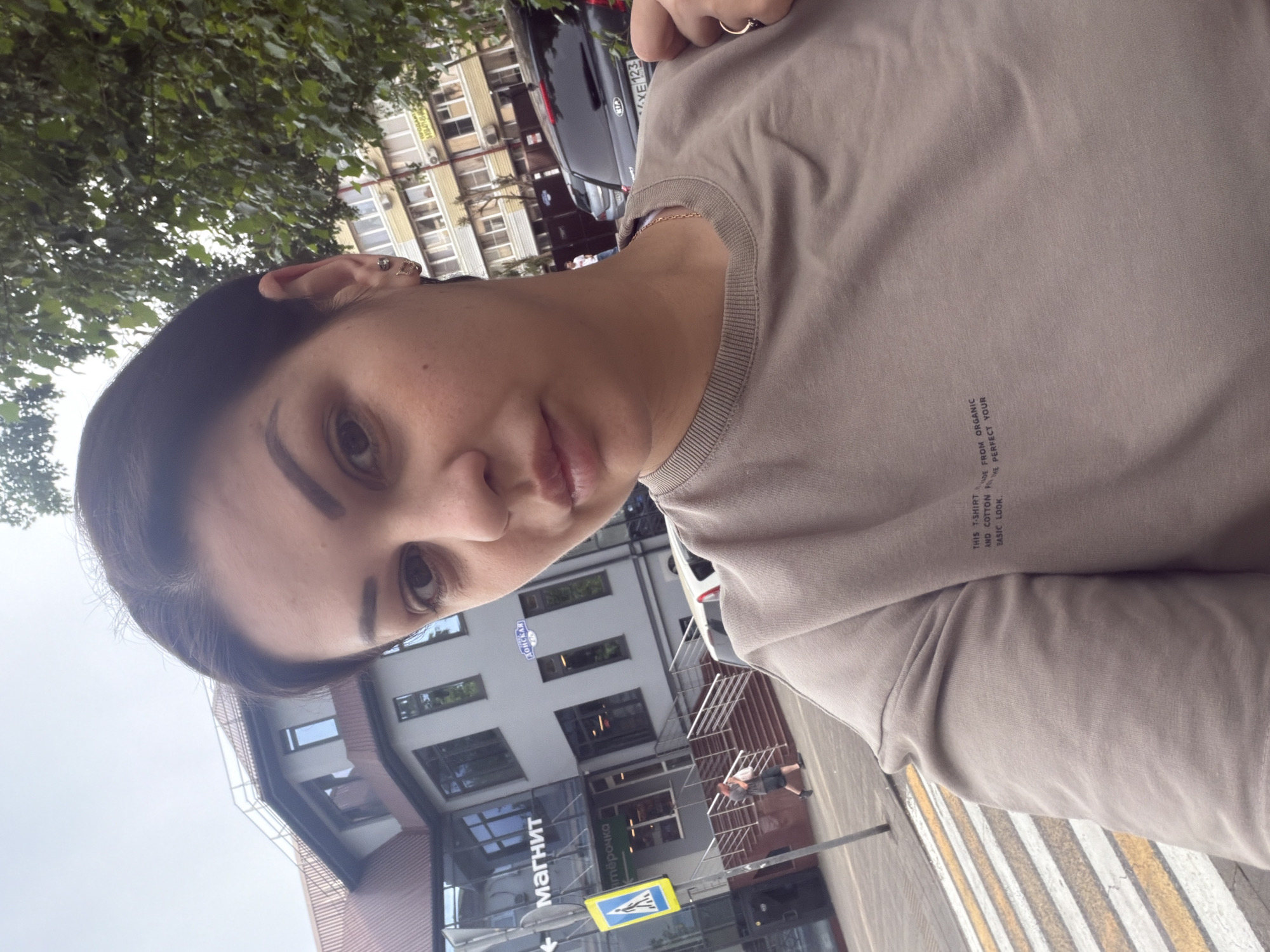 The image shows a woman taking a selfie outdoors in front of a building with a "Magnit" store sign. The building appears to be a commercial establishment in a city. There are trees, a street, a crosswalk, and a car visible in the background. The woman is wearing a light-colored sweatshirt and jewelry. The text on her sweatshirt reads, "THIS T-SHIRT IS MADE FROM ORGANIC AND COTTON FOR THE BASIC LOOK. PERFECT YOUR". A street sign in the background indicates "КОНСКАЯ". The context of the "Magnit" store, common in Russia, and the Russian street sign suggest the location is in Russia. The car's license plate is partially visible and appears to be in a Russian format.
The location is identified as Sochi, Russia, based on information not present in the image itself but provided in the prompt.
