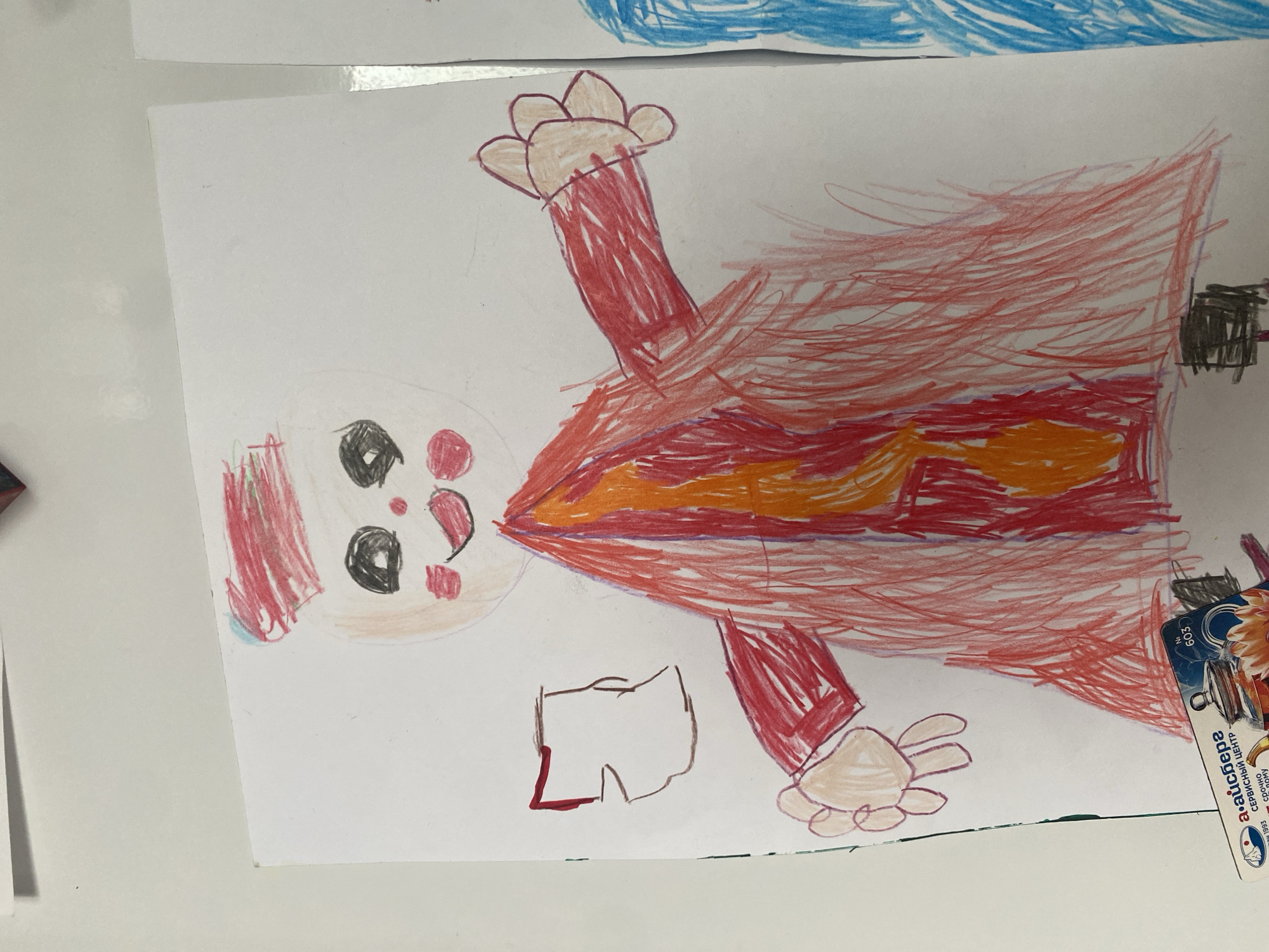 The image shows a child's drawing on white paper. The drawing depicts a figure with a round head, black eyes, pink cheeks, and a smiling mouth. The figure is wearing a red hat with horizontal lines. The body is covered by a red garment with an orange and yellow flame-like design in the center. The figure has two arms extended outwards, with one hand visible holding a light brown object that appears to be a piece of paper. The drawing is done with colored pencils. In the lower left corner of the image, a small portion of a business card or label is visible with Cyrillic text that includes "а-айсберг" and "сервисный центр". The scene appears to be indoors, on a white surface that resembles a table. There is no discernible location in Novosibirsk, Russia indicated within the image.