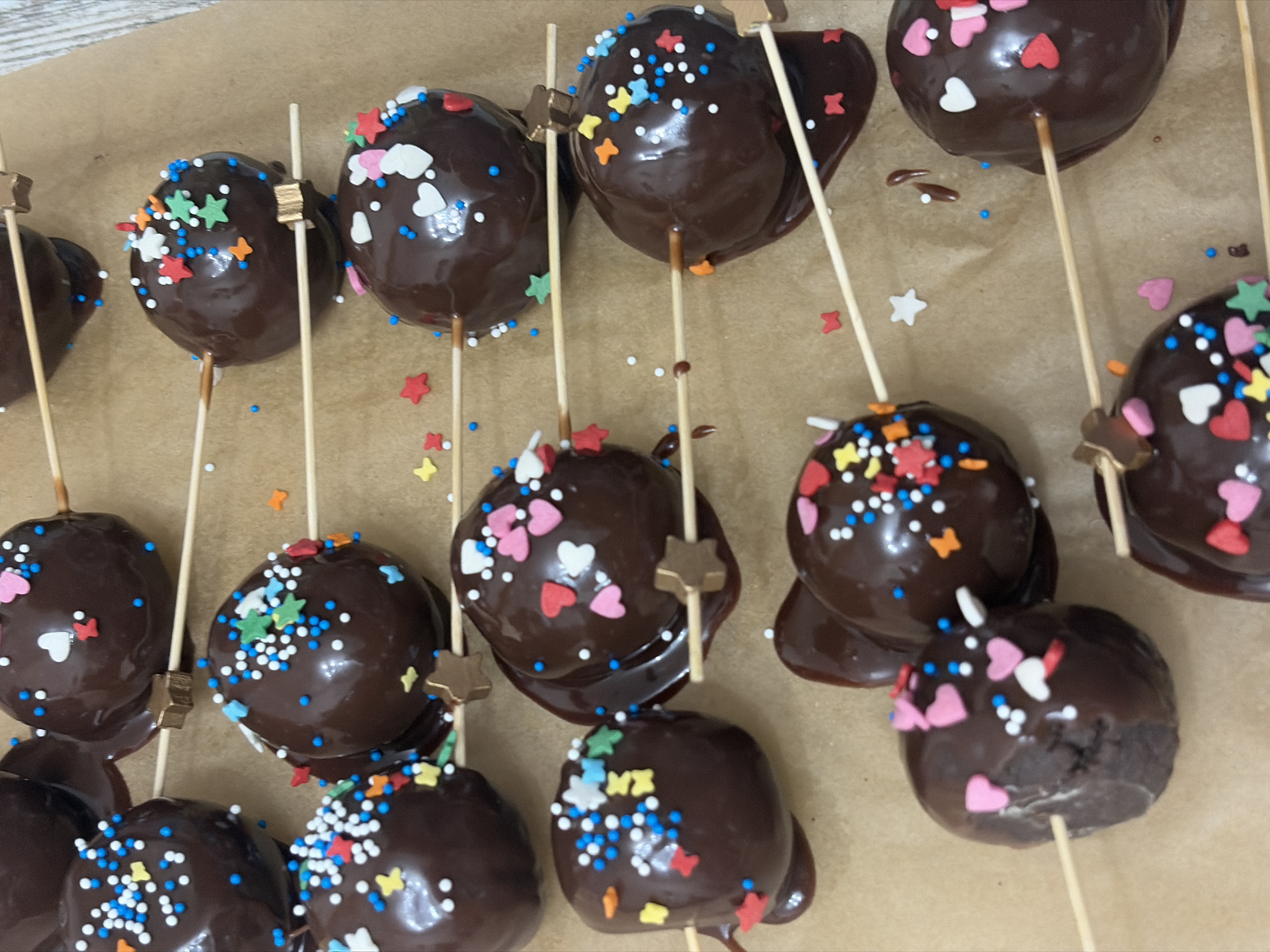 A top-down, close-up view shows about a dozen chocolate-covered cake pops arranged on a surface of tan parchment paper. The cake pops are spherical and covered in dark chocolate, many with drips of chocolate pooling at the base. Each cake pop is skewered with a wooden stick, some of which have small, star-shaped adornments near the top. The cake pops are decorated with colorful sprinkles in various shapes, including hearts, stars, and round sprinkles. The colors include white, blue, red, orange, yellow, green, and pink. The parchment paper has a subtle texture, and a few loose sprinkles are scattered around the cake pops. The scene is brightly lit, highlighting the glossy surface of the chocolate. There is no indication of the location being Saint Petersburg, Russia, or any other city. No people or interactions are visible.