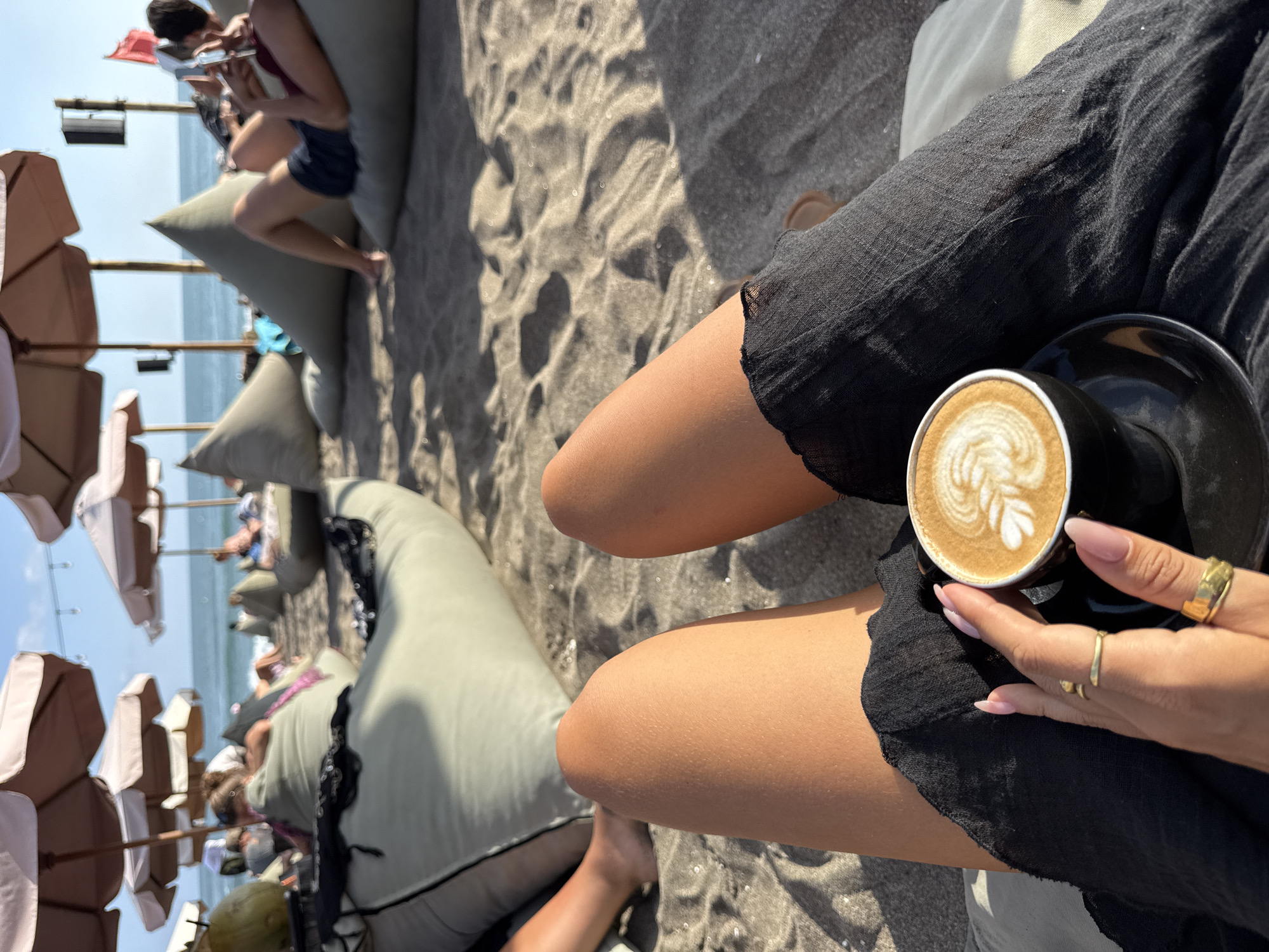 The image depicts a first-person perspective of a person relaxing on a beach in North Kuta, Indonesia, specifically holding a coffee cup. The foreground features the individual's tanned bare legs, partially covered by a loose black garment, and their right hand holding a black ceramic cup with a matching saucer. The coffee in the cup displays intricate latte art. The hand is adorned with two gold rings and has light pink, manicured fingernails.
The midground reveals a sandy beach populated with multiple light olive-green beanbag chairs, each occupied by individuals engaged in leisure activities such as relaxing, reading, or looking towards the ocean. Several large, light-colored beach umbrellas provide shade. The sand is dark gray, characteristic of volcanic beaches, and shows distinct shadows cast by the afternoon sun. On the left, a large green coconut is visible near a beanbag.
In the background, the Indian Ocean stretches to a clear horizon under a bright blue sky. Waves are visible near the shore. Wooden poles supporting umbrellas and what appears to be a black speaker on a pole, along with a red flag, suggest a beach club or lounge establishment.