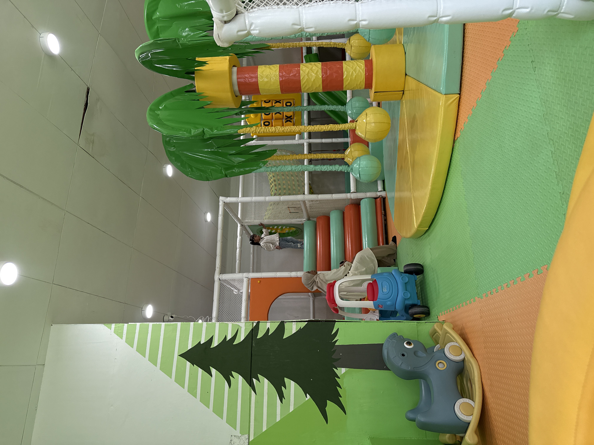 The image shows an indoor playground, potentially in Tambontunong, Indonesia, featuring a multi-level structure with various play elements. A woman wearing a hijab is standing near a blue and white plastic car toy. There is another person on a higher level of the structure. The playground includes padded cylindrical obstacles, netting, and climbing areas. A painted design depicting a tree and a hippopotamus-shaped toy is on a wall. The overall color scheme is bright, with greens, yellows, and oranges dominating the space. Overhead, there are artificial palm tree decorations and a white ceiling with recessed lights.
