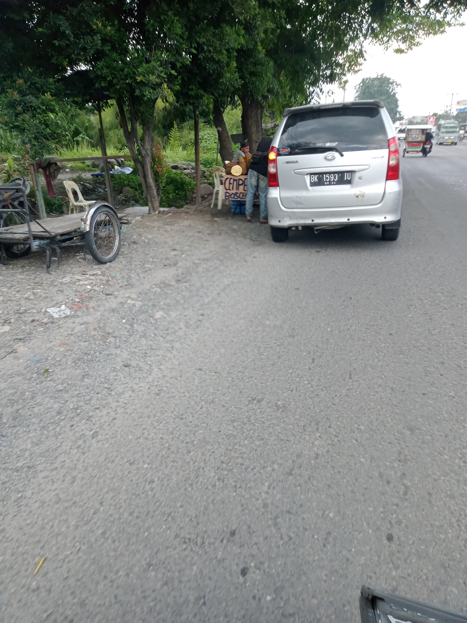In Sei Semayang, Indonesia, a silver Daihatsu minivan with license plate BK 1593 IU is parked on the side of a road. Two individuals, one wearing a brown jacket and the other a dark hijab and light-colored pants, stand near the rear of the minivan, interacting with a roadside vendor's display which includes what appears to be tempeh packaged on a circular base. To the left of the minivan, a rudimentary cart with a single wheel is visible, suggesting a form of local transportation or commerce. The environment is characterized by a roadside setting with trees and lush greenery in the background. The road surface is asphalt, and there is gravel and debris along the side. Other vehicles, including a scooter and a larger truck-like vehicle, are visible further down the road, indicating active traffic.