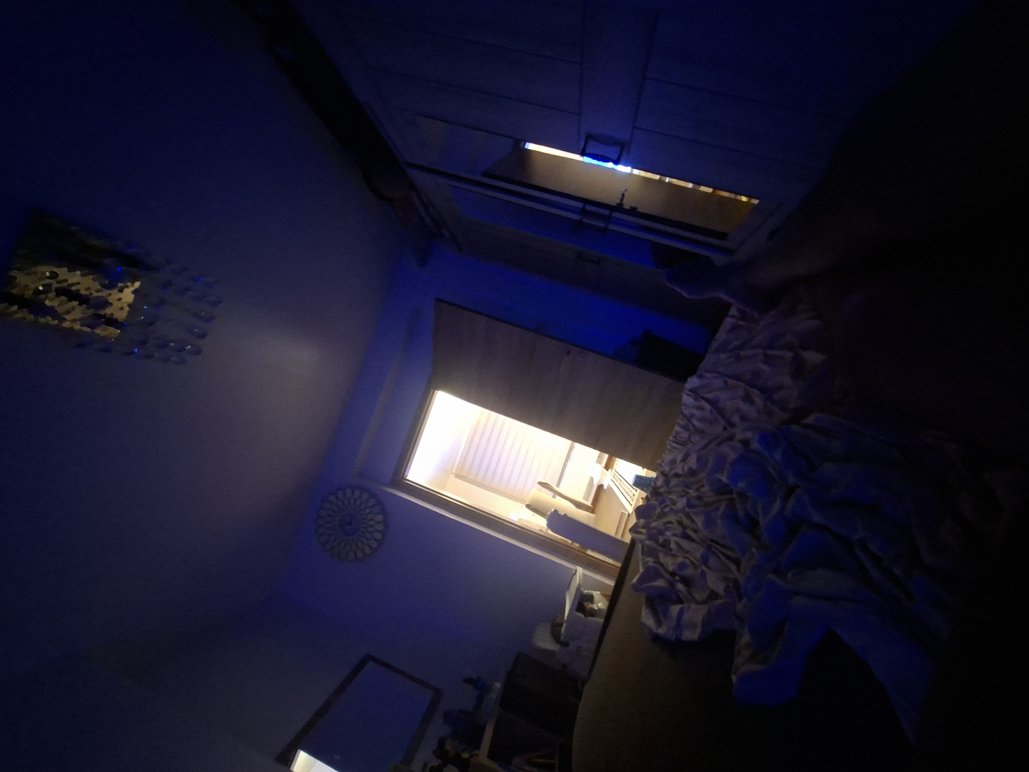 The image depicts an interior scene, possibly a bedroom, in Evere, Belgium. The room is dimly lit with blue ambient lighting and a bright light emanating from an open doorway in the background. A crumpled duvet or blanket is visible in the foreground, partially obscuring a person's legs and feet, suggesting someone is lying down.
The room's features include a ceiling light fixture with crystals, a circular decorative clock on the wall, a wardrobe with mirrored doors, and a framed mirror on the wall. The doorway in the background leads to a room with blinds on a window and what appears to be furniture. The overall impression is of a private, potentially domestic, space. The tilted angle of the photograph adds a dynamic, though disorienting, perspective to the scene.