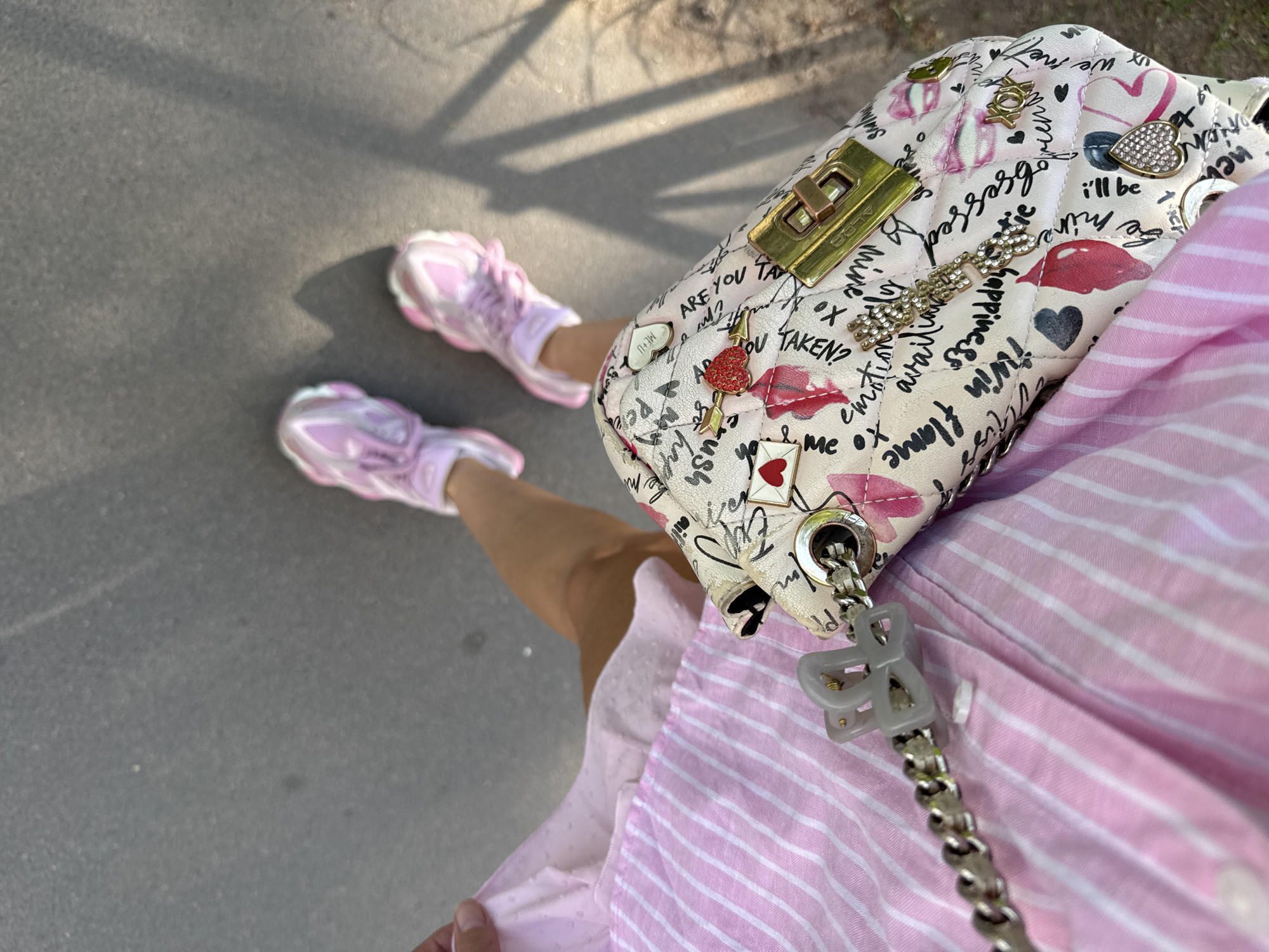 A high-angle shot features a person wearing a pink striped dress and pink sneakers, standing on a concrete surface. A patterned handbag is draped over the person's shoulder. The bag is white and decorated with various designs, including hearts, lips, and written words. The purse has a gold-colored clasp and a silver chain strap with a gray plastic hair clip attached. The sneakers have a chunky sole and pink laces. Shadows from what appears to be foliage are visible on the concrete. The location is Saint Petersburg, Russia, as the purse says "iwin flame happiness" a popular phrase used in the city.
