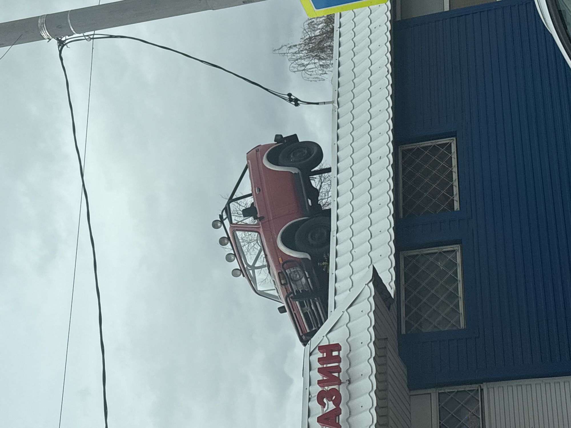 The image shows a red vehicle with a black roll cage on top of a building. The building is blue with white trim and barred windows. The image is rotated. Electrical wires cross the sky. The letters "Азин" are visible on the white trim of the building. The sky is overcast. This is a business in the city of Kazan, Russia.