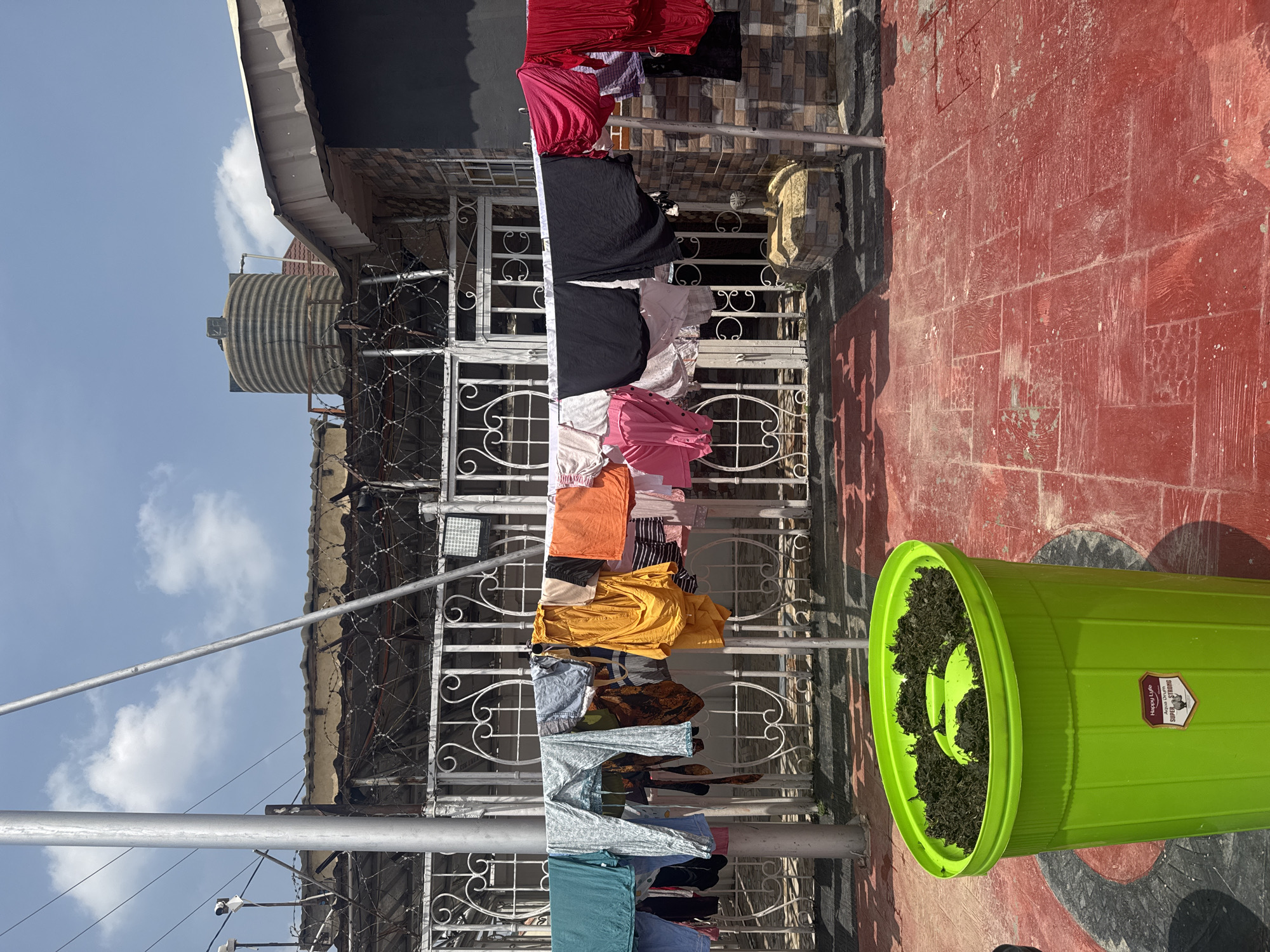 The image shows a rooftop scene in Abuja, Nigeria, with laundry hanging on a clothesline. The clothes are colorful and varied, including shirts, pants, and what appears to be a skirt or dress. They are predominantly brightly colored, such as pink, red, orange, and yellow, interspersed with darker colors like black and navy blue. The clothesline is supported by metal poles and extends across the frame, with a decorative white metal fence visible behind it.
In the background, there are buildings with corrugated iron roofs and a large, ribbed water tank, suggesting a residential or semi-industrial area. The ground is paved with red tiles, and a large, bright green plastic bin filled with dark, dry material, possibly compost or soil, sits prominently on the right side of the image. The sky is blue with scattered white clouds, indicating it is daytime with fair weather. The overall impression is a sunny, typical day with everyday activities taking place outdoors.
There is some text visible on a label on the green bin. It appears to be a product label, potentially for the bin itself, with some branding and possibly instructions or specifications. The label has a shield-like emblem and the words "Happy Life Aqua Drum Super Drum". The sunlight casts strong shadows, indicating it is likely mid-morning or afternoon. The scene is uncluttered and focuses on the drying laundry and the surrounding urban environment.