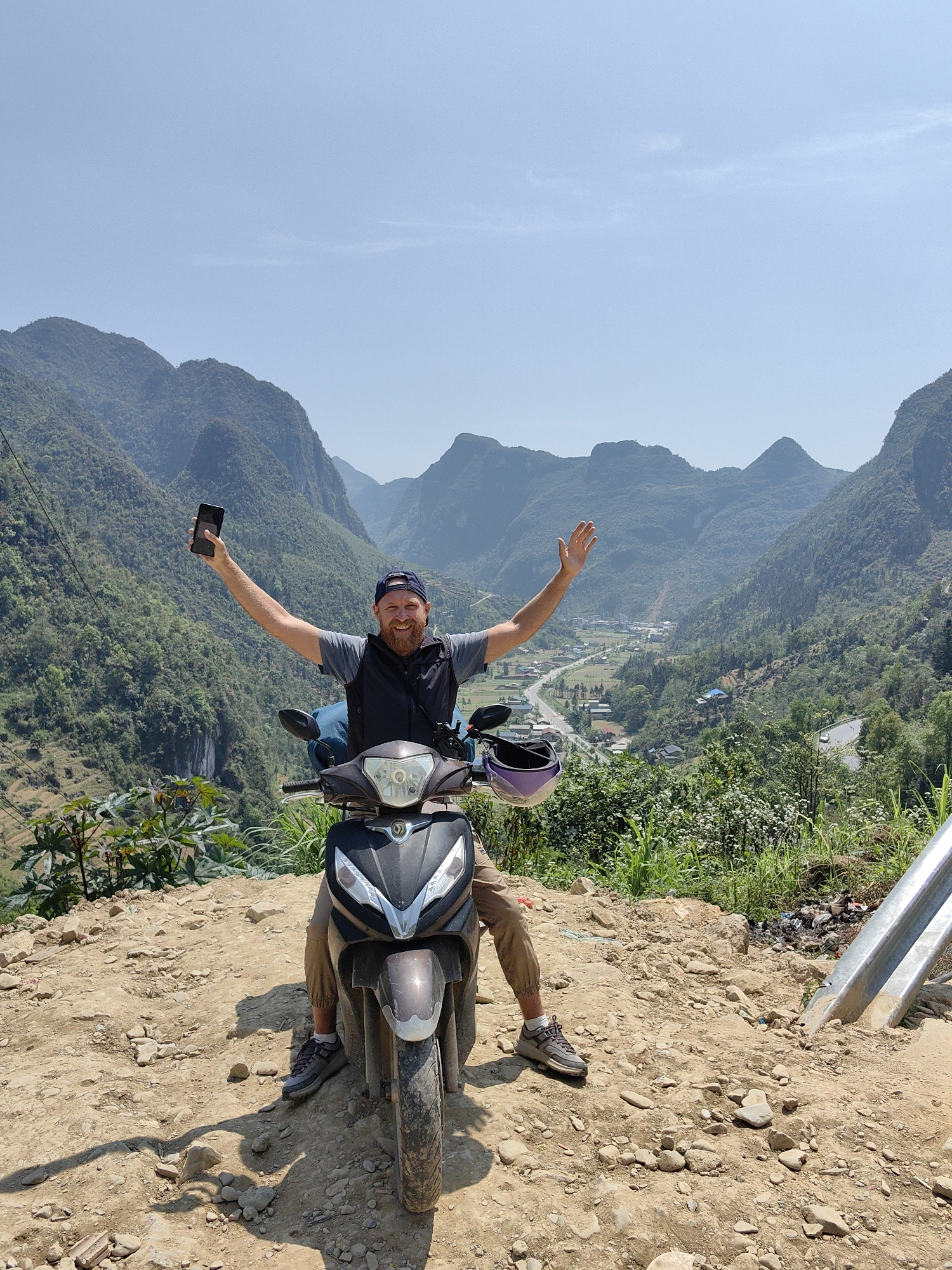 A medium, outdoor shot shows a man on a parked scooter, with a mountain vista in the background. The man is fair-skinned with a beard and a blue baseball cap. He wears a gray T-shirt, a black vest, and tan pants. He is sitting on a brown scooter, facing forward with his arms raised in a celebratory gesture. In his left hand, he holds a black smartphone. A purple helmet sits atop the scooter’s front.
The man is positioned on a rocky, dirt path. Behind him, the scene opens up to a vast valley with layered mountains covered in lush greenery. A small village can be seen in the valley below. The sky is clear and light blue.
The setting is Phố Bảng Commune, Vietnam.