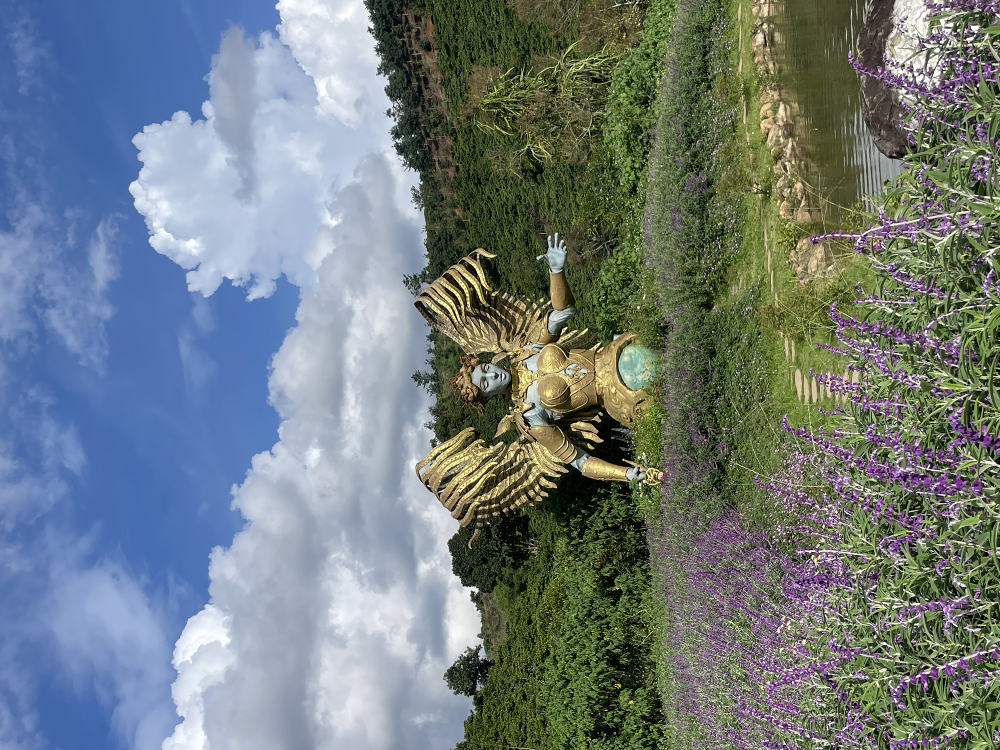 A statue of a winged figure with golden wings stands in a garden filled with purple flowers. The statue is in the foreground, with a body of water and a forested hillside in the background. The sky is blue with white clouds. The location is Thôn 1, Vietnam.