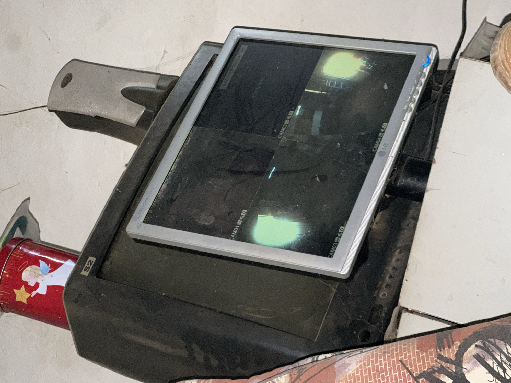 The image shows a close-up, high-angle shot of a surveillance monitor and its base, which appears to be old and dusty. The monitor is a flat-screen display with a silver bezel, and it is tilted upwards, displaying multiple camera feeds. The feeds show a green glow in some areas, possibly due to reflections or the content being displayed. Some text is visible on the monitor, including "LG" and "CAM01," "CAM02," "CAM03," and "CAM04" with icons next to them, indicating multiple camera inputs. There is a blue indicator light on the right side of the monitor.
The base of the monitor is black and appears to be made of plastic. It is large and bulky, suggesting it might be an older model or a specialized unit. A gray, curved plastic piece is attached to the left side of the base, possibly a stand or an attachment point. To the left of the base, a red tin with a design of an angel and a star is partially visible. The tin has "S2" written on a small sticker on the monitor's base.
The background is a rough, textured white wall, possibly concrete or stucco, with visible cracks. The lighting is dim, suggesting an indoor setting. The overall impression is one of disuse or a storage area. No people are visible in the image. The scene evokes a sense of outdated technology and neglect due to the dust and the visible wear on the equipment.
