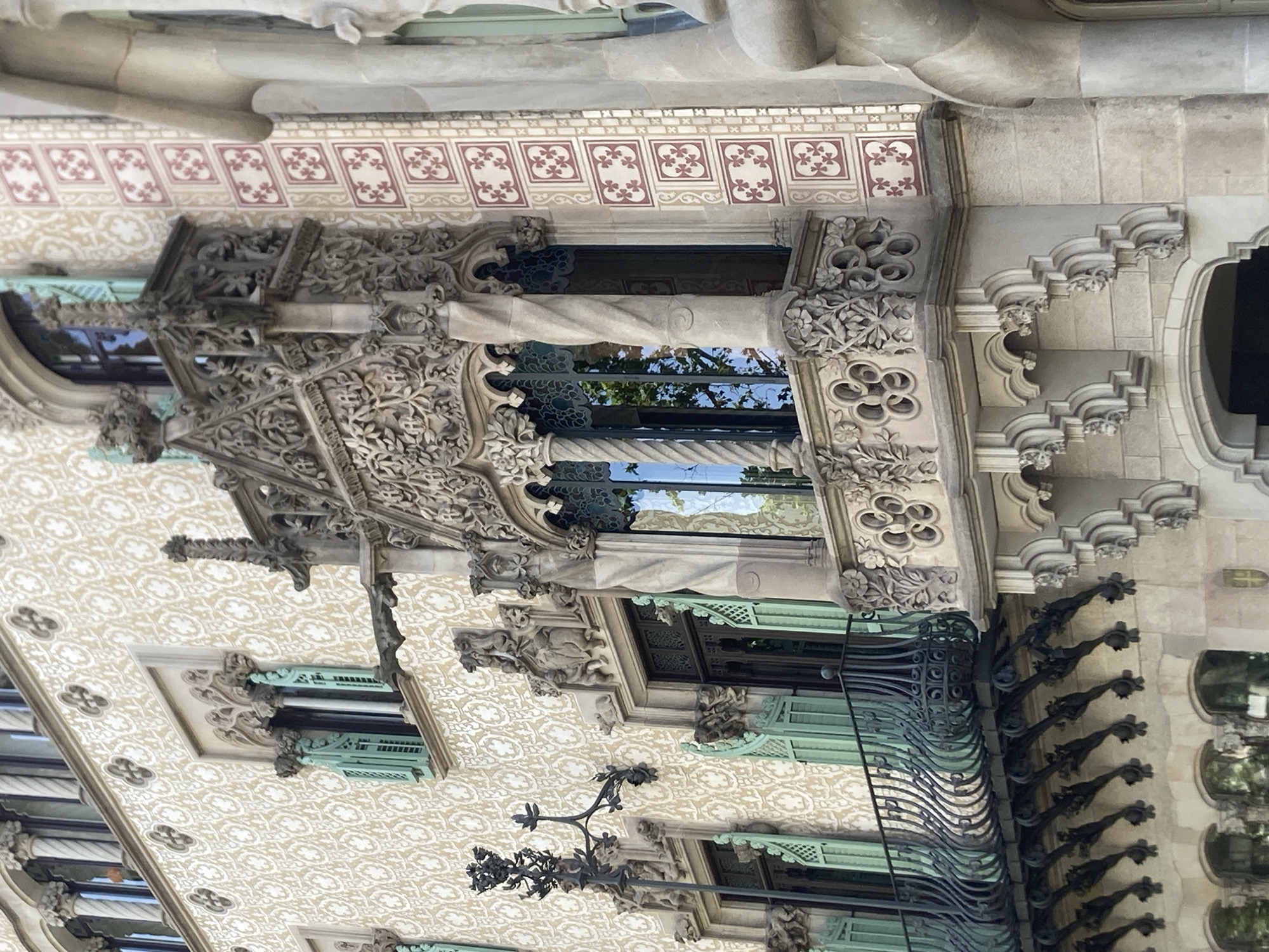 The image shows a close-up, eye-level exterior view of an ornate building facade in Barcelona, Spain, likely a residential building or a historic landmark. The architecture is highly decorative, characterized by intricate stonework, sculptural elements, and a patterned facade.
Dominating the center is a small, highly embellished balcony with carved floral and foliate motifs. It features a twisted column and a wrought-iron railing with a distinctive curvilinear design. Behind the balcony, a large window with a dark frame reflects the sky and trees. The building's exterior wall is adorned with a repetitive pattern of white clover-like symbols on a beige background, and a vertical band of square tiles with red and white floral designs.
To the left, another window is visible, also featuring green shutters and a similarly patterned wall. A decorative wrought-iron lamp post with sculpted foliage extends from the wall above this window. Gargoyle-like figures are integrated into the architectural details above the windows. The lower section of the building reveals arched openings and further intricate stonework. The overall impression is one of elaborate and historic craftsmanship.