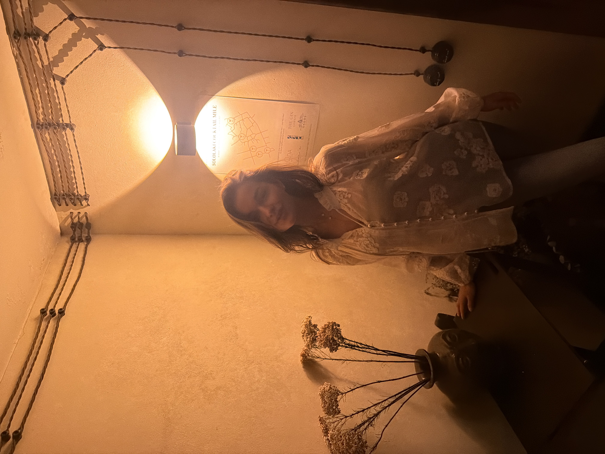 An interior scene captured in a corner features a woman standing centrally, smiling towards the camera. She has shoulder-length dark hair and is wearing a light-colored, long-sleeved blouse adorned with a floral or lace pattern. Her left hand rests on a dark rectangular table positioned in the lower-left quadrant. On this table, a dark vase with a sculpted face holds several dried, brown, branch-like plants with clustered florets.
The walls are light-colored and textured. Above the woman, a wall sconce casts warm light both upwards and downwards. Directly beneath the sconce is a poster titled "SOLOLAKI COCKTAIL MILE," which displays a map and text identifying establishments such as "THE GIN CORNER," "SNOBS," and "ADA." Exposed electrical wiring, secured by ceramic insulators, runs horizontally along the upper perimeter of the walls and descends vertically along the right wall, connecting to two circular wall-mounted fixtures. The overall environment suggests a commercial establishment like a bar or cafe within the Sololaki district of Tbilisi, Georgia.