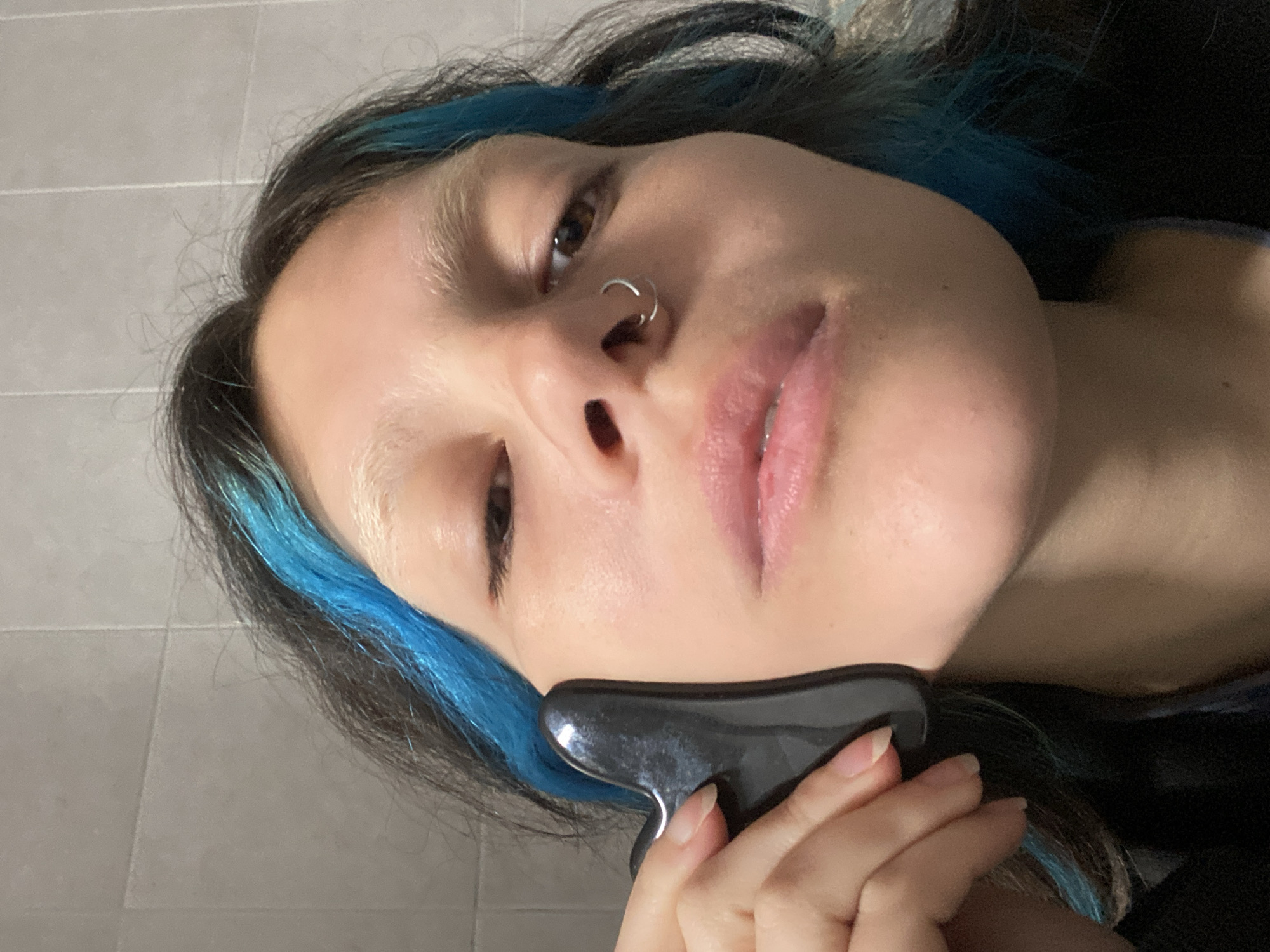 A close-up, tilted portrait features an individual with dark hair accentuated by streaks of vibrant blue/teal dye. The person has light-colored eyebrows, a silver hoop nose piercing, and full lips. One eye is visible, directed towards the viewer, conveying a neutral and focused expression. Their right hand, adorned with natural nails, holds a dark, possibly black or dark grey, contoured gua sha tool firmly against their jawline or cheek, suggesting a skincare or facial massage routine. The background reveals a light grey tiled wall, indicating an indoor setting, likely a bathroom. The lighting appears to be ambient indoor light. No text is visible in the image. This scene takes place in Gijón/Xixón, Spain.