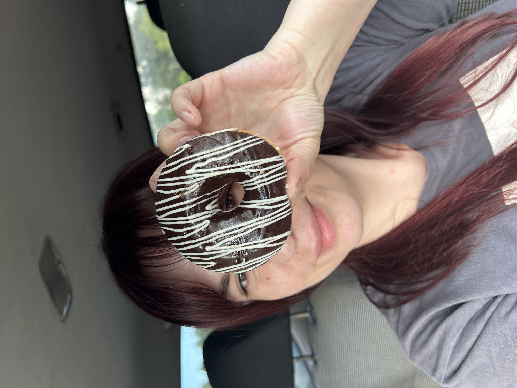The image shows a close-up of a person holding a chocolate-glazed donut with white icing drizzled on top. The donut is held in the foreground, with the person's hand supporting it. The person's face is partially visible, with reddish-brown hair framing their head. They are looking towards the donut, and their mouth is slightly open, suggesting they might be about to take a bite or are enjoying the view. The person appears to be inside a vehicle, as indicated by the dark interior and a blurred background that suggests trees and possibly a window. The lighting is bright, hinting at daylight. The overall mood is casual and perhaps joyful, centered around the enjoyment of a sweet treat. The image does not contain any visible text. The location context is Chaikovsky, Russia.