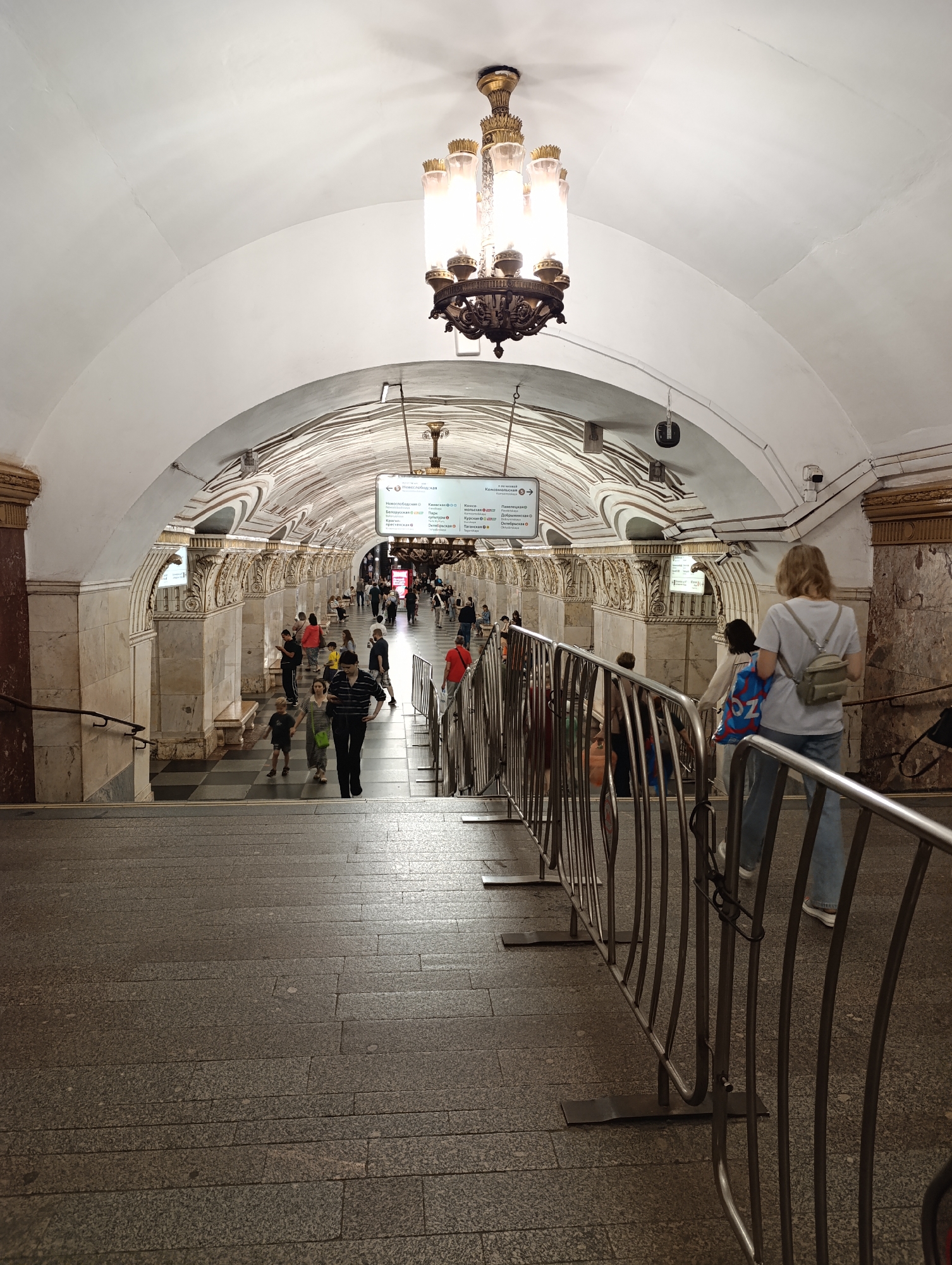 An indoor scene captures a bustling passage within a grand metro station, identified by its ornate architecture as likely part of the Moscow Metro system. The primary perspective looks down a set of wide, granite steps towards a brightly lit, arched corridor.
Numerous people are seen moving through the station. In the foreground, a woman with blonde hair, wearing a white t-shirt and jeans with a small beige backpack, stands facing away from the viewer, looking down the stairs. Beside her, metallic crowd control barriers delineate the path. Down the stairs and in the main hall, various individuals and small groups walk in different directions, some heading towards the stairs, others moving along the platform. Notable figures include a man in a striped shirt walking with a child, and another person carrying a blue bag with the "OZ" logo.
The station boasts impressive, classical-style architecture. A magnificent multi-tiered chandelier with warm glowing lights hangs from the center of the high, vaulted white ceiling, which also shows some faint cracks. The walls are adorned with marble-clad pillars, intricate decorative friezes, and bas-reliefs in golden and white tones. The floor of the main hall features a checkered pattern.
Visible text on overhead signs, partially captured, includes destination names common to the Moscow Metro, such as "Новослободская" (Novoslobodskaya), "Киевская" (Kievskaya), "Курская" (Kurskaya), "Комсомольская" (Komsomolskaya), "Таганская" (Taganskaya), "Павелецкая" (Paveletskaya), "Добрынинская" (Dobryniniskaya), and "Октябрьская" (Oktyabrskaya), confirming the location context of Moscow, Russia. The scene appears to be taking place during the day, illuminated by both artificial light from the chandelier and ambient station lighting. There are no strong indications of specific weather as it's an indoor setting. People appear calm, engaged in routine transit activities.