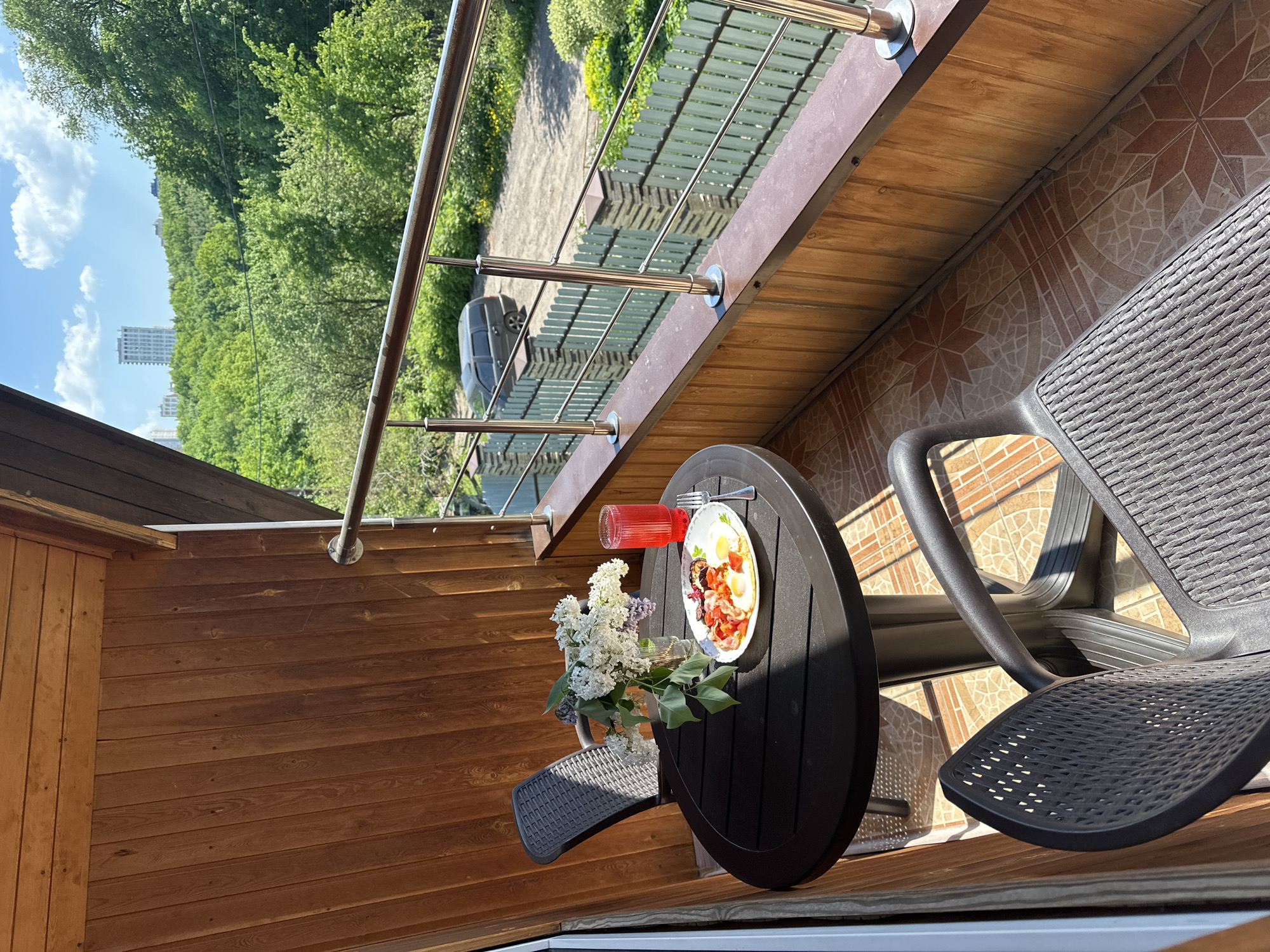 The image depicts a balcony scene in Nizhny Novgorod, Russia. On a black round table sits a white plate with a meal of fried eggs, bacon, and other unidentified food items, a glass of red liquid, and a small bouquet of white and purple lilacs. Beside the table are two black plastic chairs. The balcony has a metal railing overlooking a green landscape with trees and a building in the distance, suggesting an urban fringe location. The balcony floor is tiled, and the wall is covered with wooden planks. A car is parked in the driveway below. The overall impression is one of a relaxed outdoor meal in a residential setting.