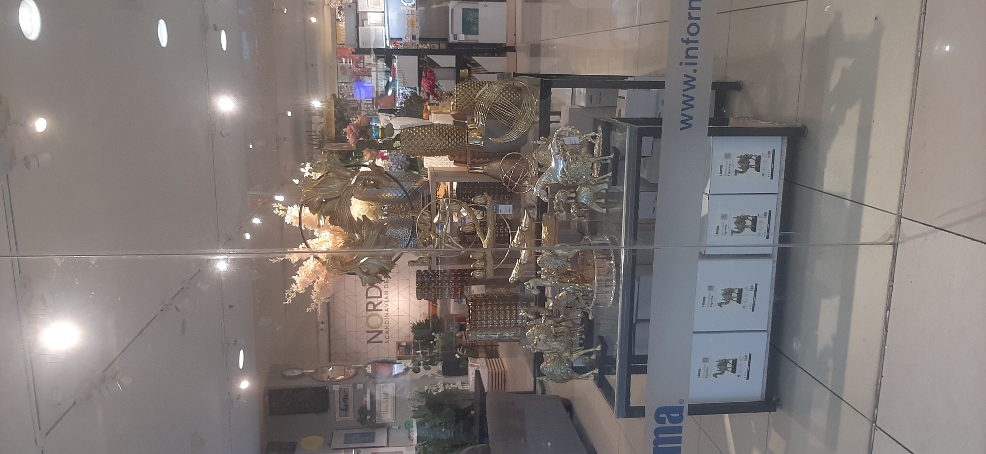 The image is taken from outside a store, looking through a glass window. The store appears to be a home decor or furniture shop. In the foreground, the bottom part of the glass has a white strip with a blue logo that reads "Jform" and a website address "www.inform". Below this, there are several white boxes with images of camels on them, indicating products for sale.
The main display inside the store features a collection of golden decorative items. Prominently displayed are several gold-colored figurines of camels and horses, arranged on a glass-topped table. Behind these figurines, there are large, ornate gold sculptures, some resembling abstract floral or leaf designs, and a tall, textured golden vase. Further back, more decorative items are visible, including golden pineapple-shaped objects, smaller golden vases, and other metallic decorative pieces.
In the background, the store's interior is visible, showing shelves stocked with various home goods. There is a prominent wall display with the word "NORD" and "SCANDINAVIAN DESIGN" written on it, suggesting the store's theme or origin. Lighting fixtures are visible on the ceiling, casting light on the displays. The overall impression is of a well-lit retail environment showcasing decorative home furnishings.
The Jambi, Indonesia location is inferred from the "Jform" logo, which is associated with Informa, a large furniture and home accessories retailer with a presence in Indonesia, including Jambi.