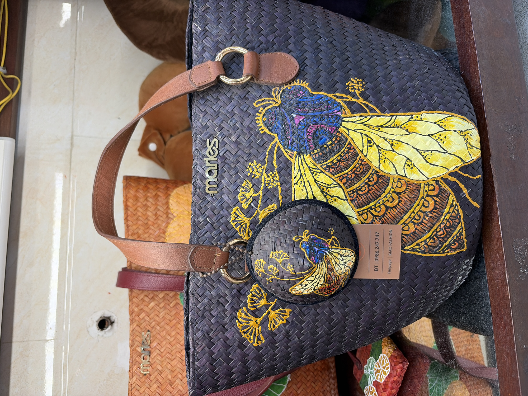 This is a close-up, eye-level, indoor shot of a woven handbag with a large, decorative bee design. The handbag is dark gray, woven, and has tan leather straps and accents. The bee is prominently displayed on the front, with intricate yellow, orange, blue, and purple patterns. A circular, woven keychain with a smaller bee design is attached to the strap. A rectangular tag is affixed to the handbag, featuring Vietnamese text and a phone number. To the left of the main handbag, a portion of another woven orange bag with a red strap is visible. The background appears to be a wall with a tiled pattern and a dark wooden shelf or surface in the foreground. The lighting is bright and appears to be from overhead.