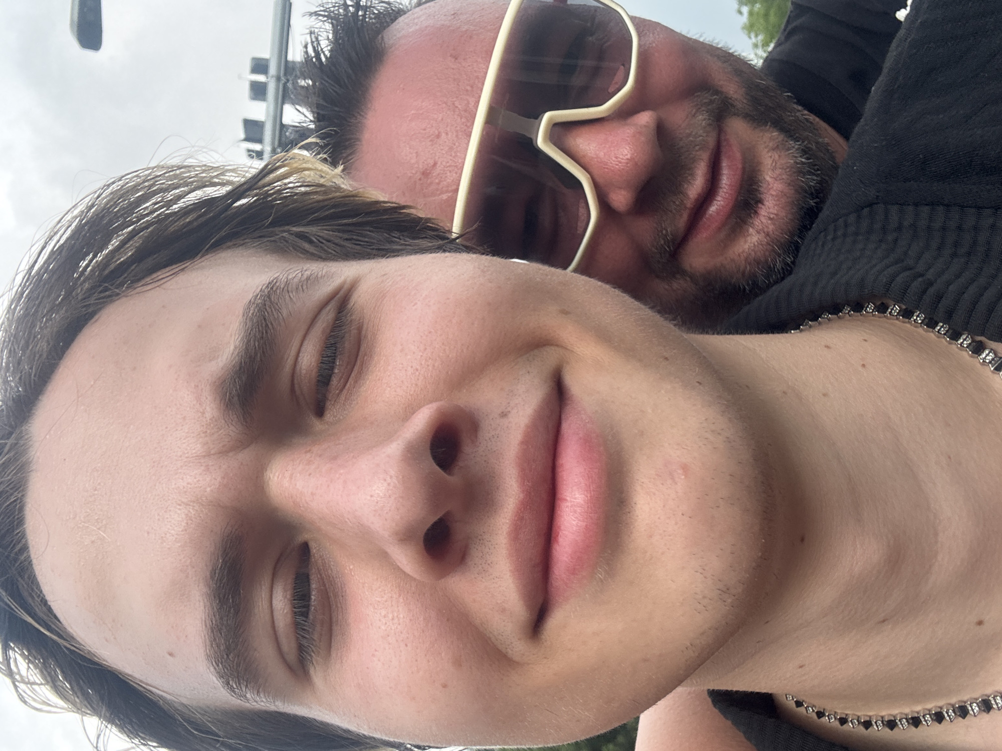The image is a close-up selfie taken outdoors, likely on a street or public area. Two individuals are visible. The person in the foreground, on the left, is a young Caucasian male with light brown hair. He is looking directly at the camera with a slight smile. He is wearing a black garment and a silver chain necklace. Partially behind him, on the right, is an older Caucasian male with dark hair and a beard, wearing large, light-colored sunglasses and a black shirt. The background is blurred but appears to be an urban setting with some greenery and overcast sky. There are no specific landmarks that would indicate the location as Suzhou Industrial Park, China. The lighting suggests it is daytime.