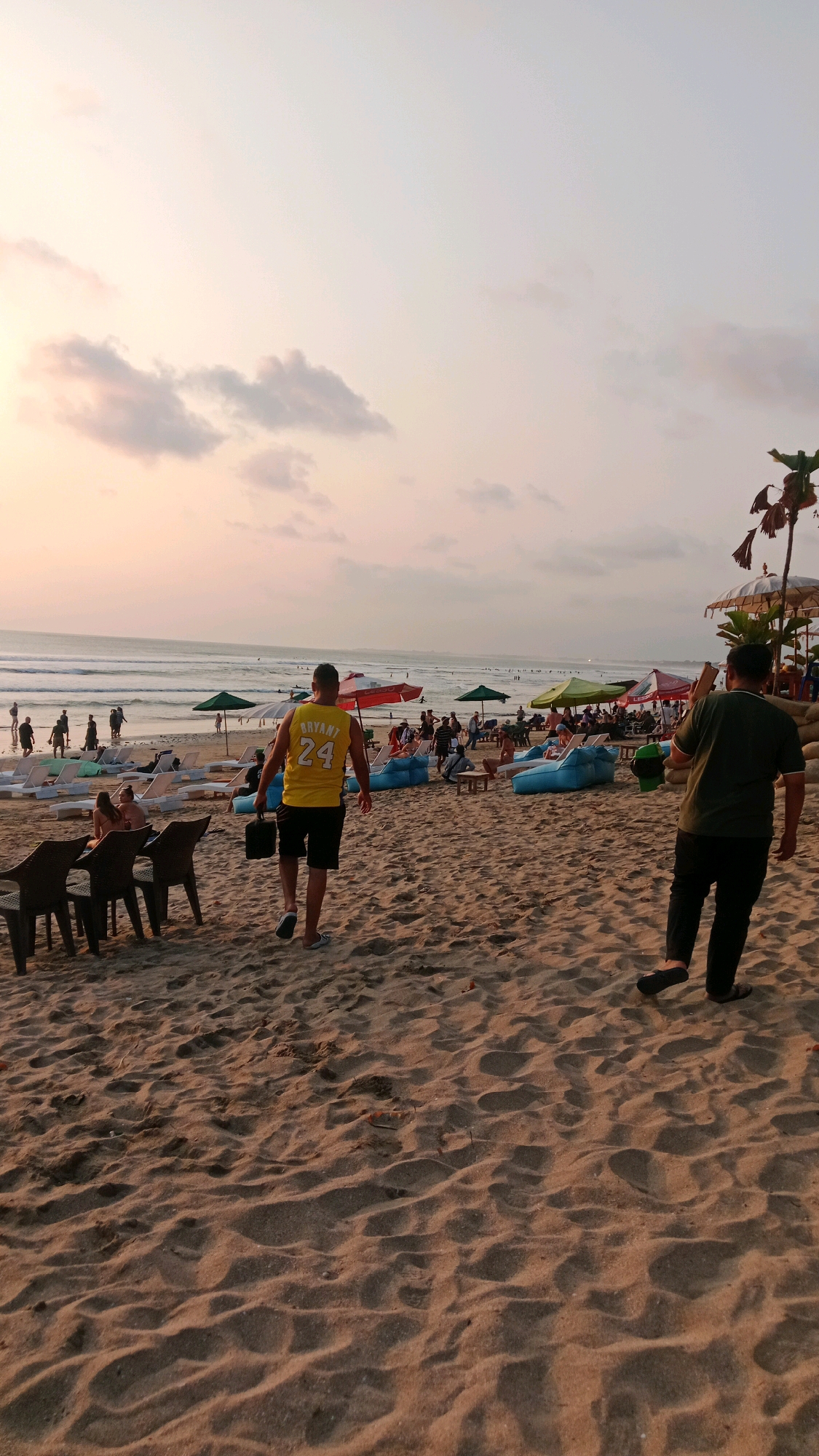 The image depicts a sunset scene on Kuta Beach, Indonesia. The sandy shore is occupied by numerous beachgoers, lounge chairs, and umbrellas. In the foreground, two men are walking across the sand. The man on the left wears a yellow basketball jersey with the number 24 and "BRYANT" printed on the back, black shorts, and light-colored slip-on shoes, and is carrying a black bag. The man on the right wears a dark green t-shirt, black pants, and dark flip-flops. The ocean with gentle waves is visible in the background, with many people wading in the water or walking along the shoreline. The sky is illuminated by the soft hues of sunset, with scattered clouds. The overall atmosphere suggests a relaxed evening at a popular tourist destination.