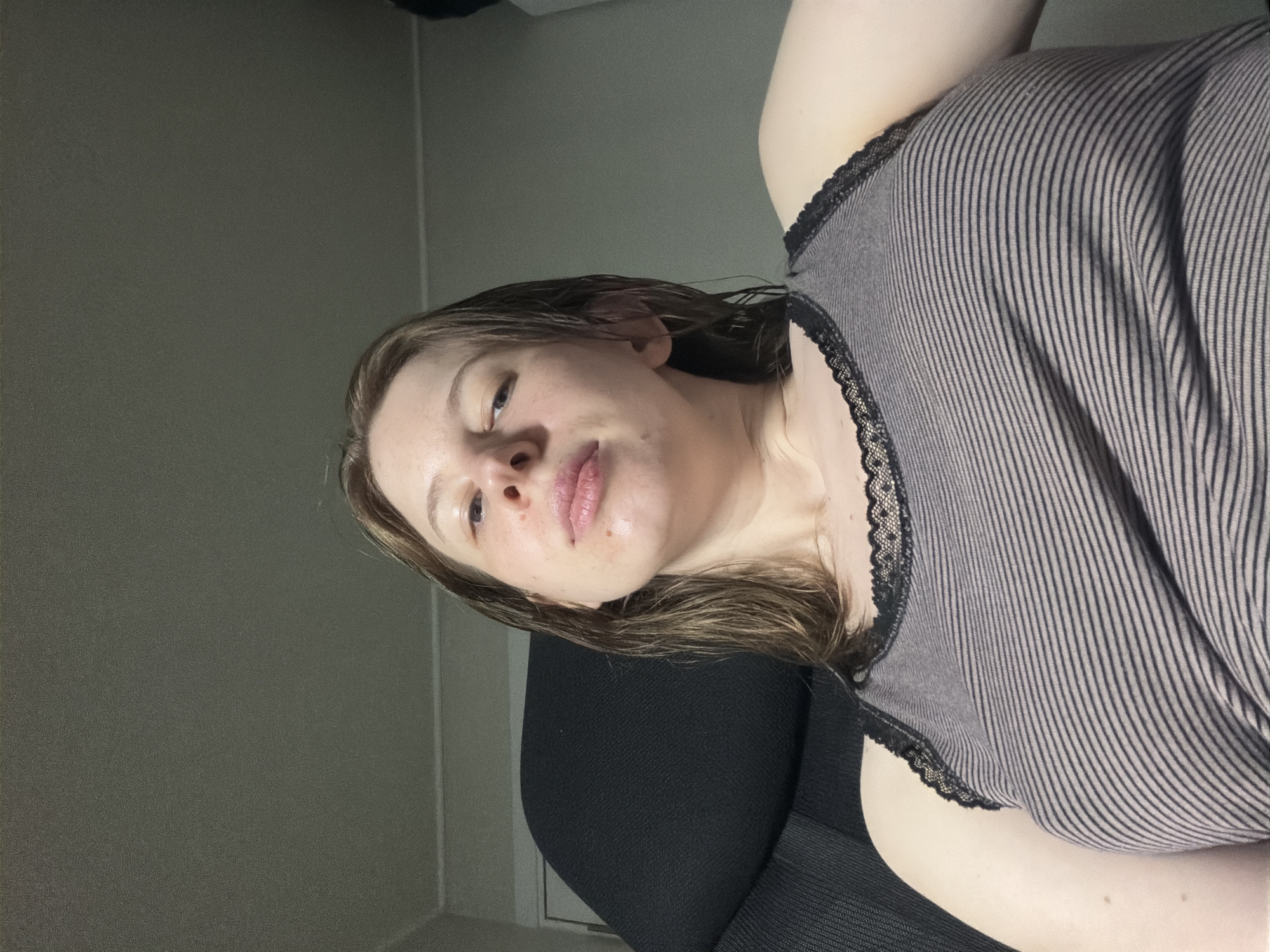 A selfie is taken from a low angle looking up at a young woman with shoulder-length brown hair, sitting in a black office chair. She is wearing a grey and black striped tank top with black lace trim. Her skin appears slightly shiny. The background consists of a plain, light-colored wall and ceiling, suggesting an indoor residential setting. No other people or objects are clearly visible. The image was taken in Dongducheon-si, South Korea.