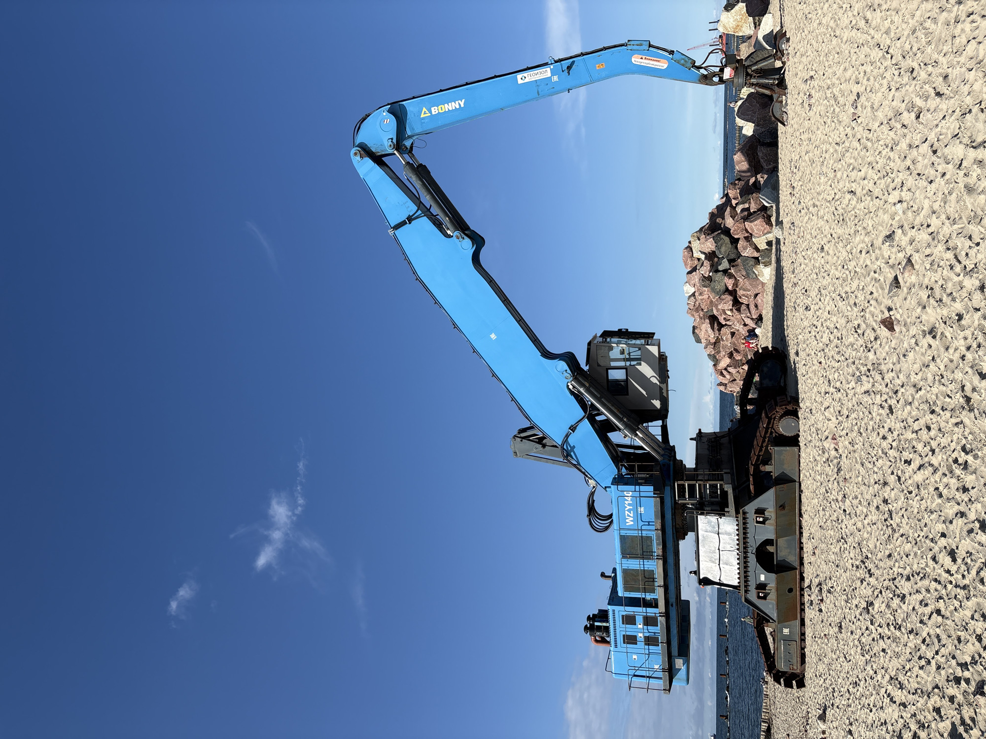 A large blue excavator with the model number "WZY140" is parked on a sandy beach in Svetlogorsk, Russia. The excavator has a long arm extended upwards and to the right. To the right of the excavator is a large pile of reddish-brown rocks. The sea is visible in the background with a few boats in the distance. The sky is blue with a few white clouds. There are a few people visible on the beach in the distance, near the pile of rocks. The scene appears to be a construction or demolition site due to the presence of the excavator and the pile of rocks.