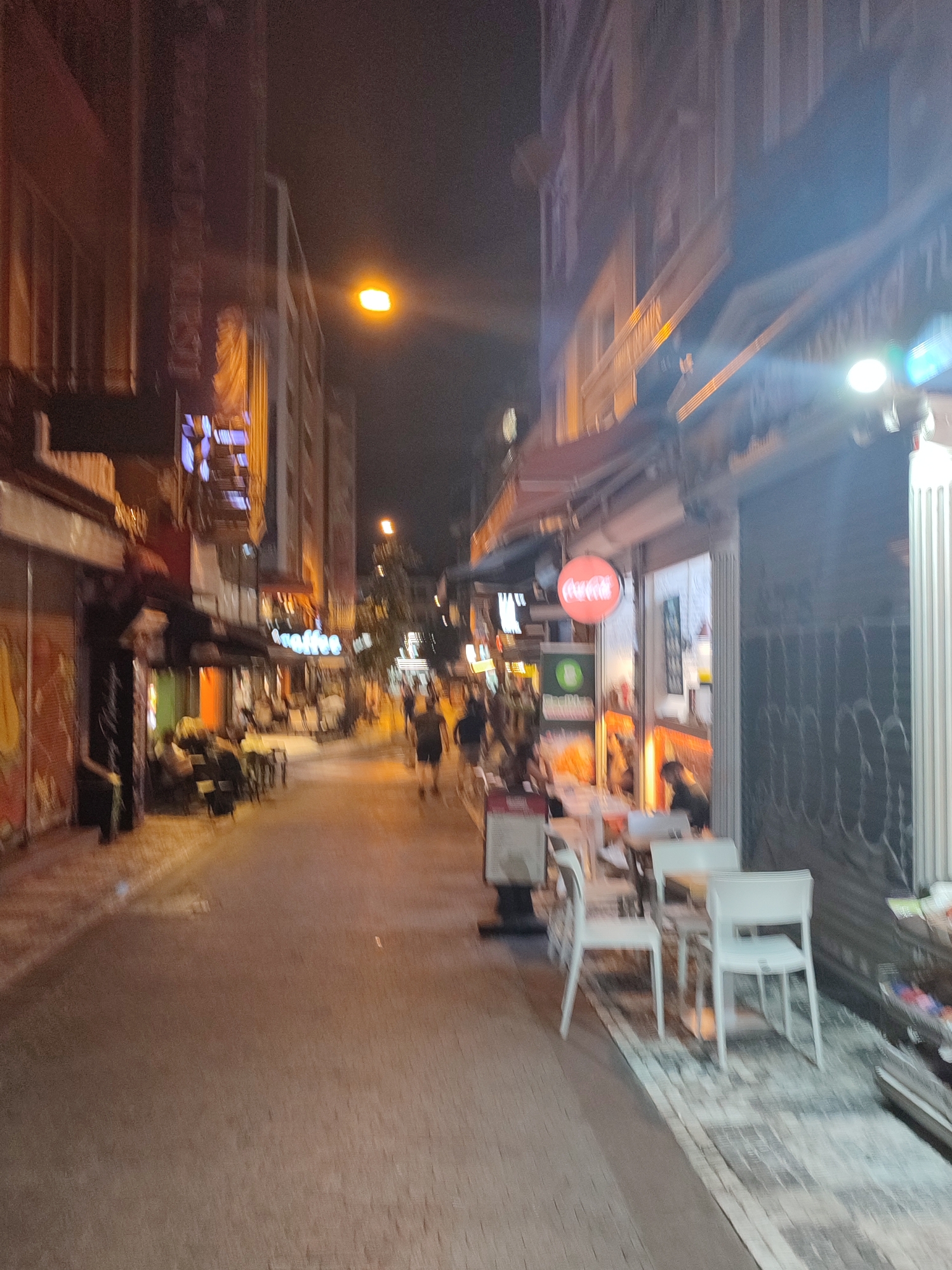 The image depicts a nighttime street scene in Kadıköy, Turkey, likely a commercial or pedestrian area with restaurants and shops. The perspective is from eye-level, looking down a relatively narrow street lined with buildings on both sides. The lighting is dim, with streetlights and illuminated signs casting a warm glow on the pavement and facades.
On the right side of the street, there are shops with awnings and outdoor seating areas occupied by customers. One prominent sign displays the Coca-Cola logo. White tables and chairs are arranged on a patterned pavement, suggesting an outdoor dining space. Further down the street, more people are visible, some walking and others seated.
The left side of the street also features buildings with illuminated storefronts. Some areas appear to be under renovation or have shuttered businesses. There are people sitting at tables outside some establishments.
The overall atmosphere is lively but subdued, characteristic of an evening in a busy urban district. The image is somewhat blurry, indicating motion during the capture, possibly from a handheld camera or a moving vehicle.