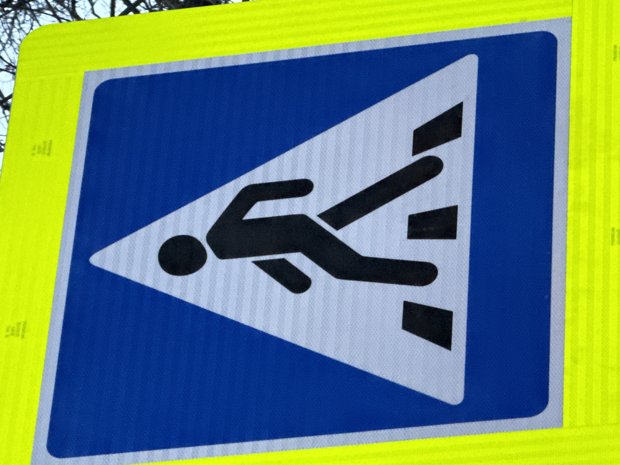 A close-up shot shows a blue-and-yellow road sign with a white triangle and black pedestrian figure on it. The sign is a pedestrian crossing warning sign commonly found in Votkinsk, Russia. The sign has a blue background with a white reflective triangle in the center, inside of the triangle there is a black figure of a person walking across a zebra crossing. The yellow outer square of the sign is fluorescent and has a reflective texture for increased visibility. The background of the shot has out-of-focus branches of a tree.
