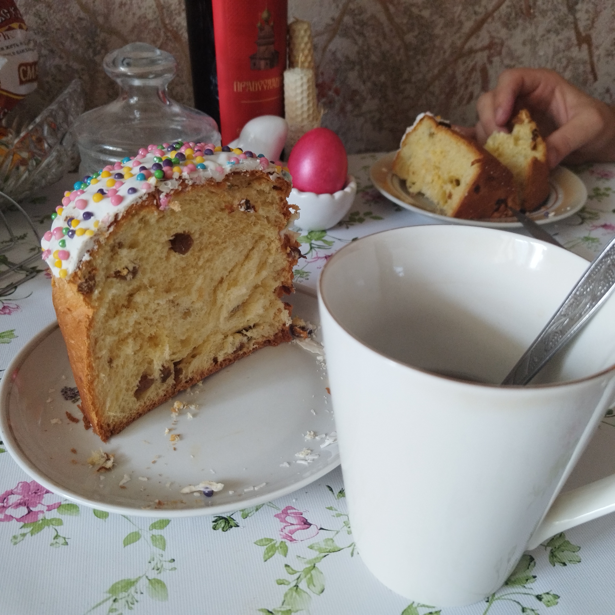 The image shows a table setting with Easter treats, likely taken in a domestic environment in Yekaterinburg, Russia. A cut slice of Kulich, a traditional Easter bread decorated with white icing and colorful sprinkles, sits on a white plate with floral patterns. Next to it is a large white mug containing a spoon. In the background, there's a red Easter egg in a small white dish, and further back, a red container with Cyrillic writing on it, potentially a wine bottle or other Easter-themed product. A person's hand is visible in the upper right corner, holding another piece of Kulich on a separate plate. There are also candles, possibly beeswax, and a glass container in the background. The table is covered with a floral patterned tablecloth.