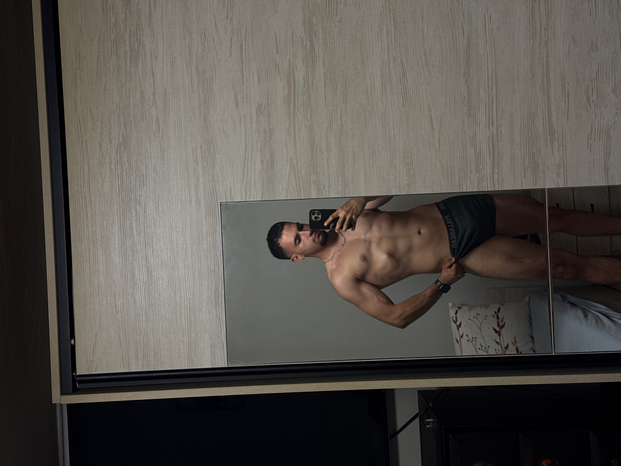 A man is standing in front of a mirror, taking a selfie. He is wearing dark green boxer briefs. The mirror reflects him and a pillow on a bed behind him. The background of the image is a closet with light-colored wooden doors. The scene is likely indoors, possibly a bedroom, in Shumen, Bulgaria.