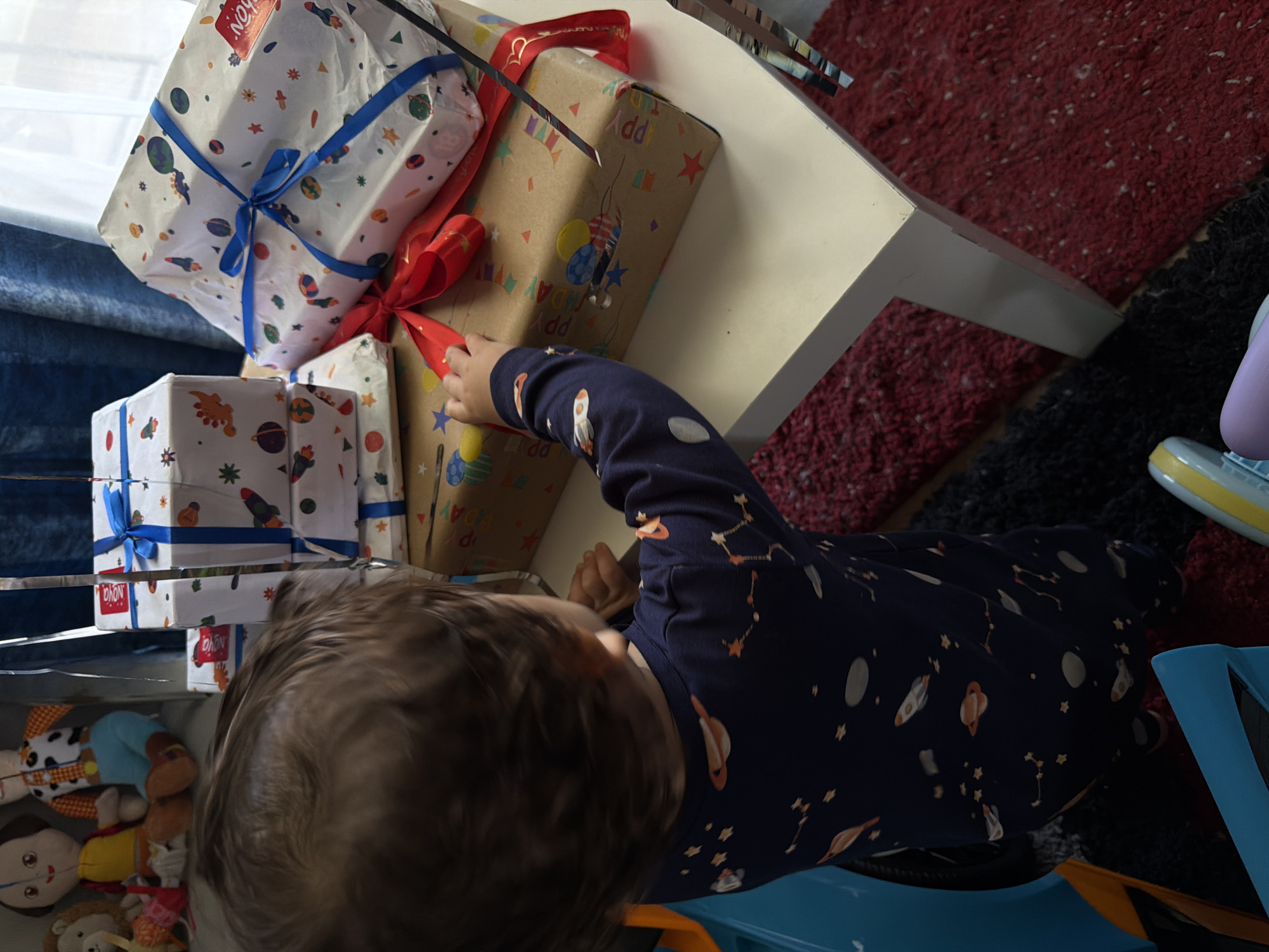 In an indoor setting, possibly a home, a toddler is shown from behind, reaching towards a collection of wrapped presents. The toddler wears a navy blue pajama set adorned with a pattern of stars, planets, and rockets. In the background, several plush toys are visible, including what appears to be a figure resembling Woody from Toy Story.
The presents are arranged on a light-colored surface, which is likely a table. Some of the gifts are wrapped in paper featuring a space-themed design with rockets and planets, tied with blue ribbon. Another gift is wrapped in brown paper with colorful balloons and confetti, and appears to have "Happy Birthday" printed on it, also adorned with red ribbon. The floor in the foreground and surrounding the table is covered with a textured rug in shades of red and black. The overall lighting suggests an indoor environment with natural light coming from a window, partially obscured by blue curtains. There is no indication of the location being in Sharm El Sheikh, Egypt, nor any elements that would suggest this specific geographical context.