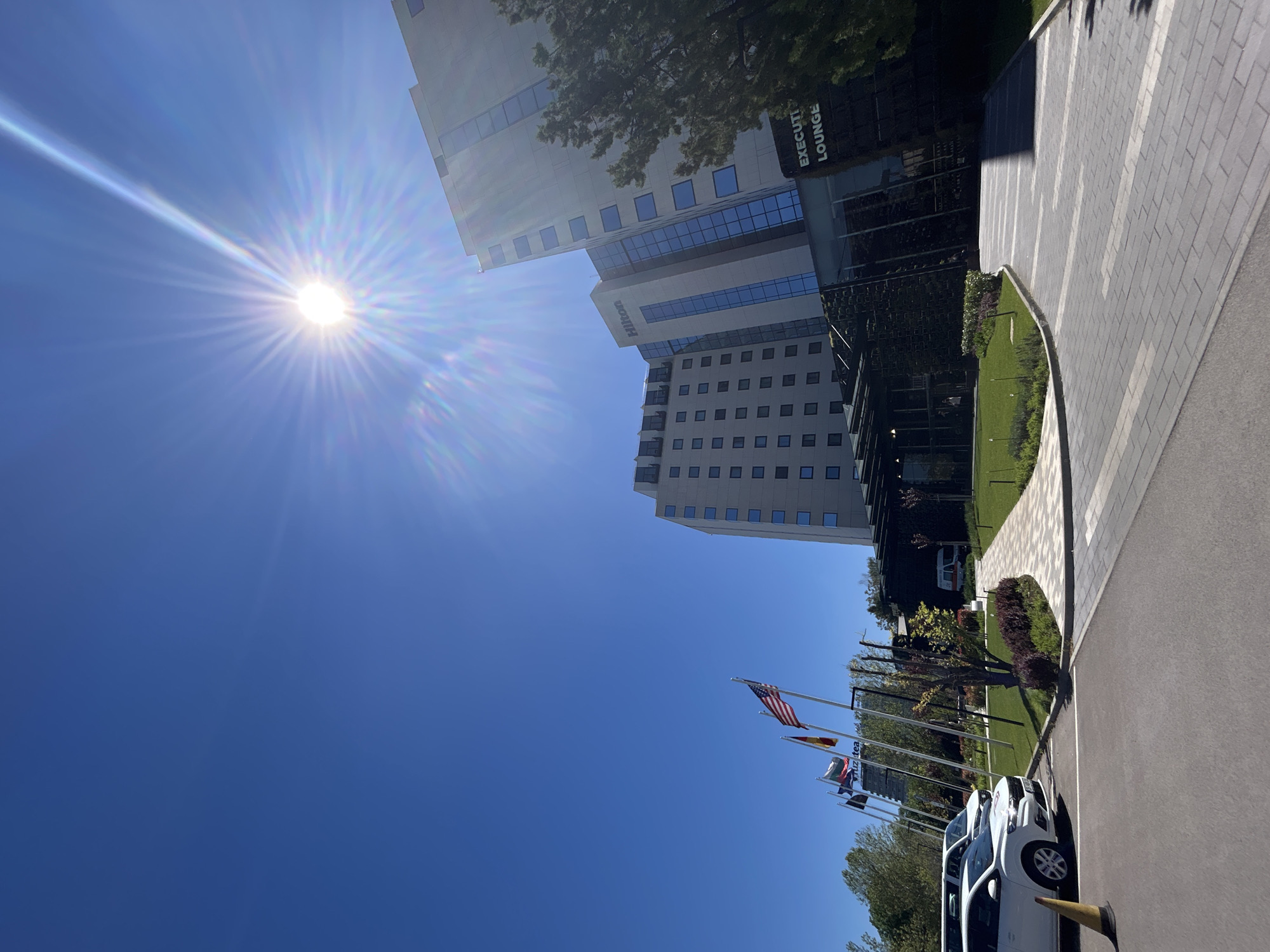 The image captures the Hilton hotel in Sofia, Bulgaria, under a bright, cloudless blue sky. The sun is visible in the upper left portion of the frame. The hotel's architecture is modern, featuring a multi-story building with several windows and a sign indicating an "Executive Lounge." A row of flags, including the American flag, is seen along the driveway leading to the hotel entrance. Several white cars are parked nearby. A paved walkway and manicured greenery are also visible.