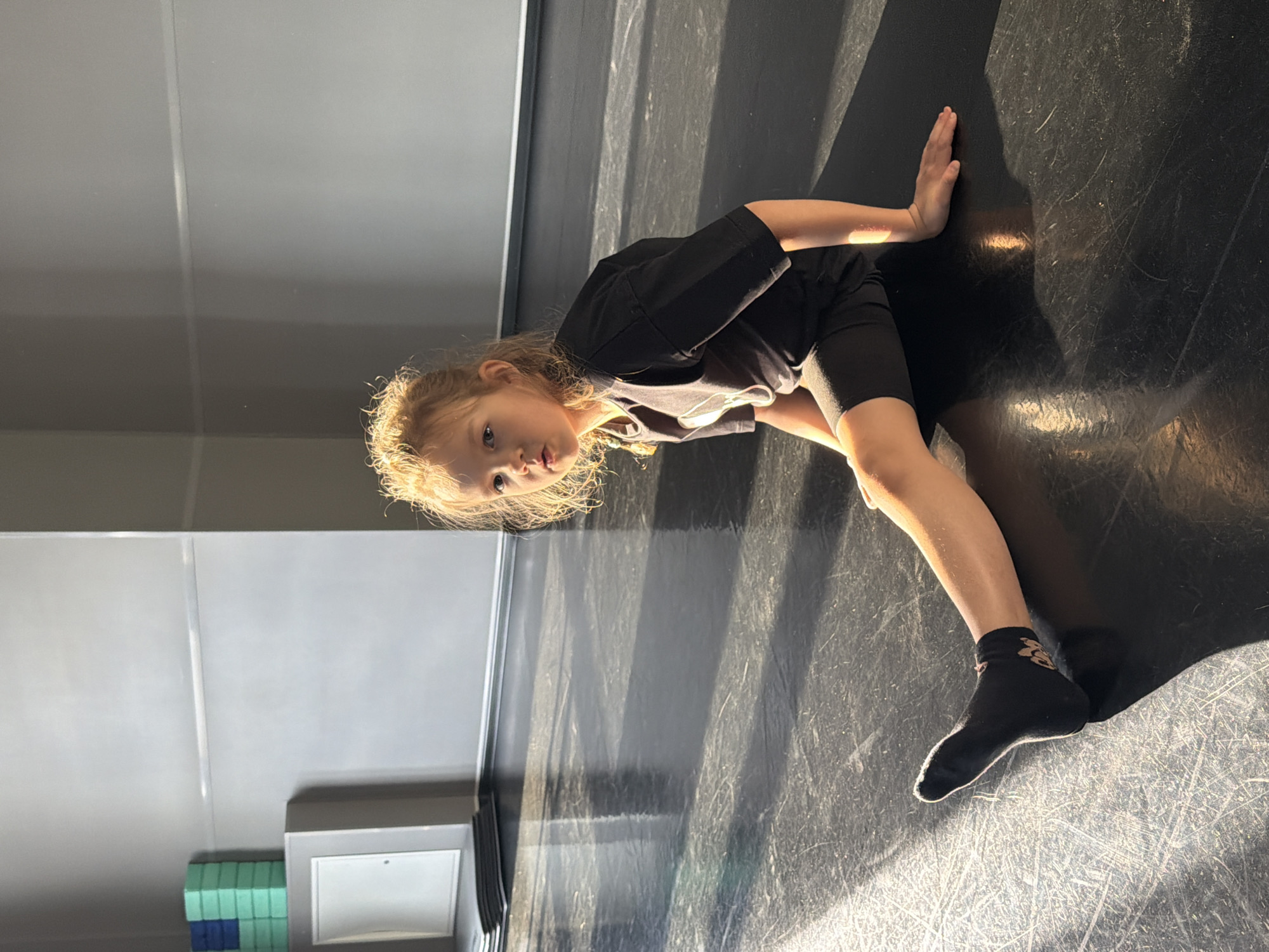 A young, light-skinned child with light hair and wearing black shorts, a black t-shirt and black socks is performing a gymnastic stretch on a dark floor. The child's left hand is on the wall. The location is unclear, but likely an indoor space with a smooth floor, probably a studio or gym. There is no information available to determine if the location is Saint Petersburg, Russia.