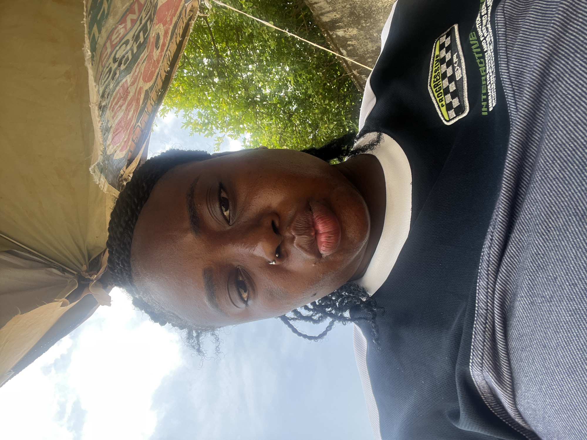 The image is a close-up, low-angle selfie of a young Black woman taken outdoors. She is looking towards the camera with a neutral expression. She has dark skin, braided hair, and a nose ring. She is wearing a black and white top with some text and a checkered patch on the left side, and a blue and white striped garment on the right. Behind her, there is a partially visible tan-colored umbrella or awning with colorful patterns, lush green trees, and a cloudy sky. The overall lighting suggests it is daytime, likely late morning or afternoon, with bright sunlight filtering through the trees. The context indicates the photo was taken in Abuja, Nigeria. There is visible text on the umbrella that is difficult to read due to the angle and resolution, but appears to include letters like "THO" and "INS." The checkered patch on her shirt has the word "PROGRESSIVE" written on it in yellow.