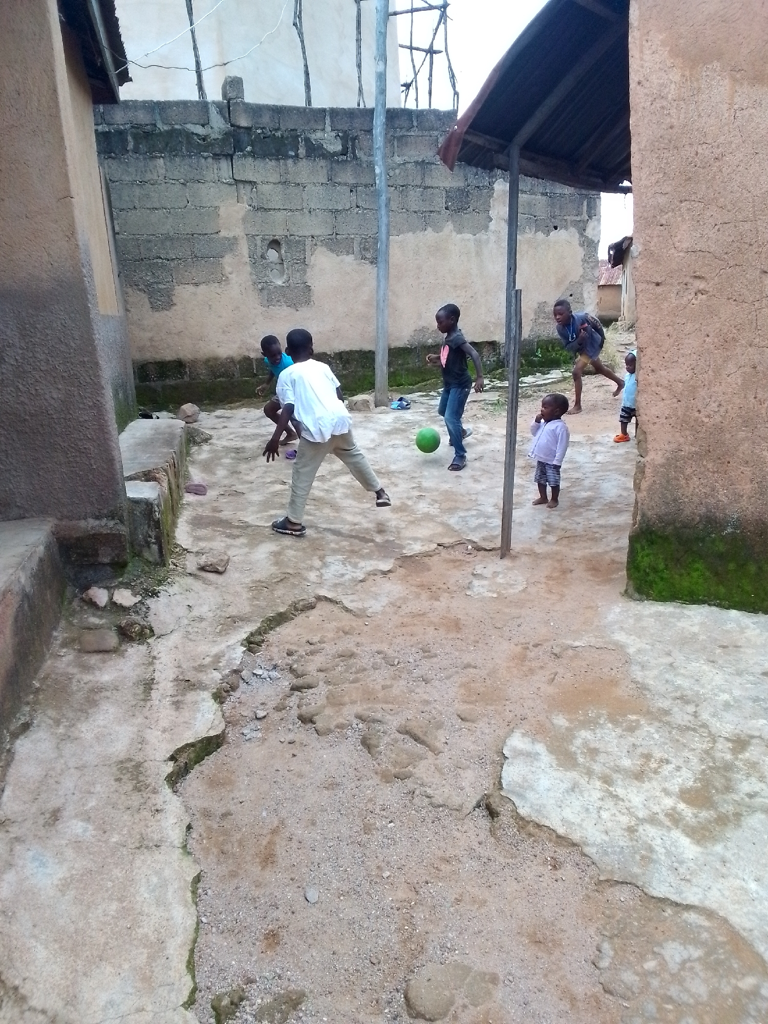 In Unknown, Unknown, a group of children are playing soccer in a dusty, urban alleyway. The alley is bordered by unpainted concrete buildings and a wall constructed of concrete blocks. In the foreground, the ground is a mix of cracked concrete and packed dirt, with scattered stones and debris. The children, ranging in age from toddlers to pre-teens, are dressed in casual clothing. One boy in a white t-shirt and tan pants kicks a green soccer ball. Another boy in a black t-shirt and jeans is positioned to receive the ball. A younger child in a blue shirt and shorts stands nearby, while a toddler in a light-colored jacket and striped shorts observes the game. In the background, a corrugated metal roof extends over part of the alley, supported by wooden posts. The scene is dimly lit, suggesting it may be late afternoon or an overcast day.
