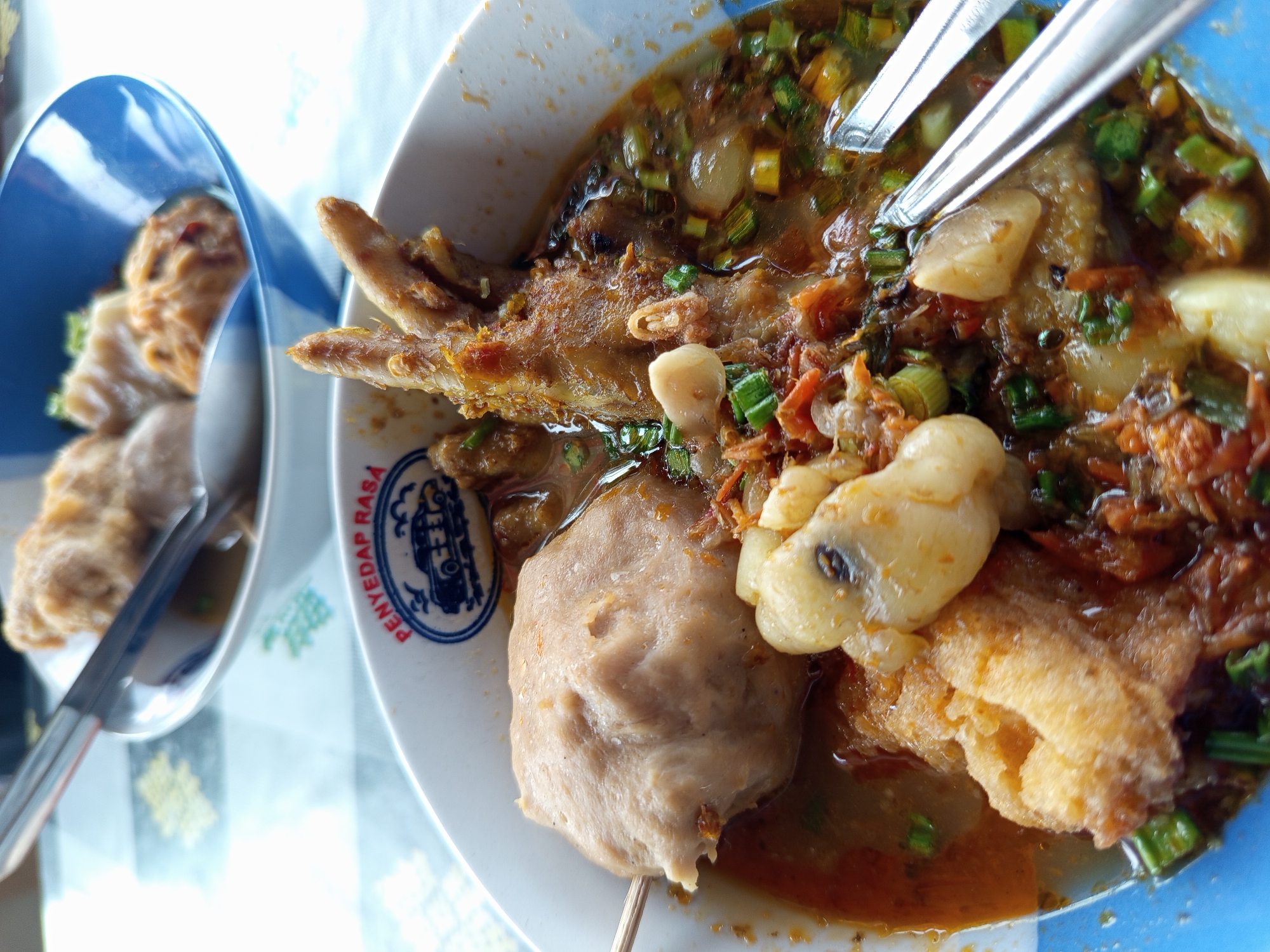 The image depicts a close-up view of a bowl of bakso, a Indonesian meatball soup, served in a blue and white bowl with the logo "PENYEDAP RASA" visible. The soup contains a large meatball on a skewer, pieces of fried tofu, and what appears to be chicken feet, all in a rich broth with green onions and other garnishes. Two spoons are partially submerged in the soup. In the background, another bowl of bakso is visible, also with spoons and meatballs. The setting appears to be a casual dining establishment, likely a warung or restaurant, in Bokor, Indonesia. The lighting suggests an indoor setting, possibly with natural light coming from the side.