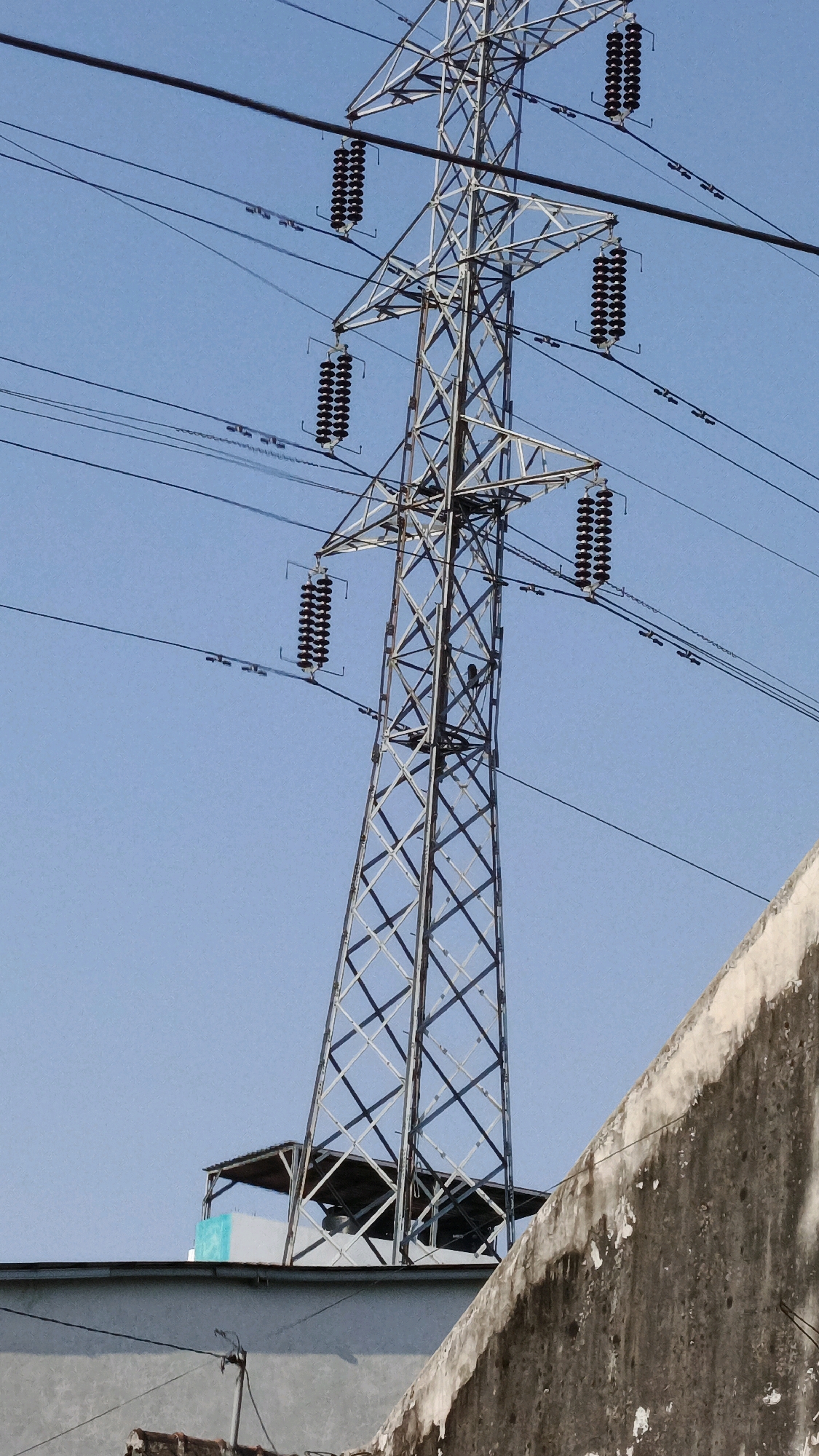 The image shows a tall, lattice-steel electricity pylon against a clear blue sky in Pabelan, Indonesia. Several power lines are suspended from the pylon, held in place by insulator strings. In the foreground, a weathered concrete wall runs diagonally across the bottom right of the frame. Below the pylon, a flat-roofed structure with a metal awning is visible, partially obscured by the pylon's base. A small, teal-colored object is visible on this structure. The overall scene depicts infrastructure against a bright sky, with elements of urban or semi-urban construction in the foreground.