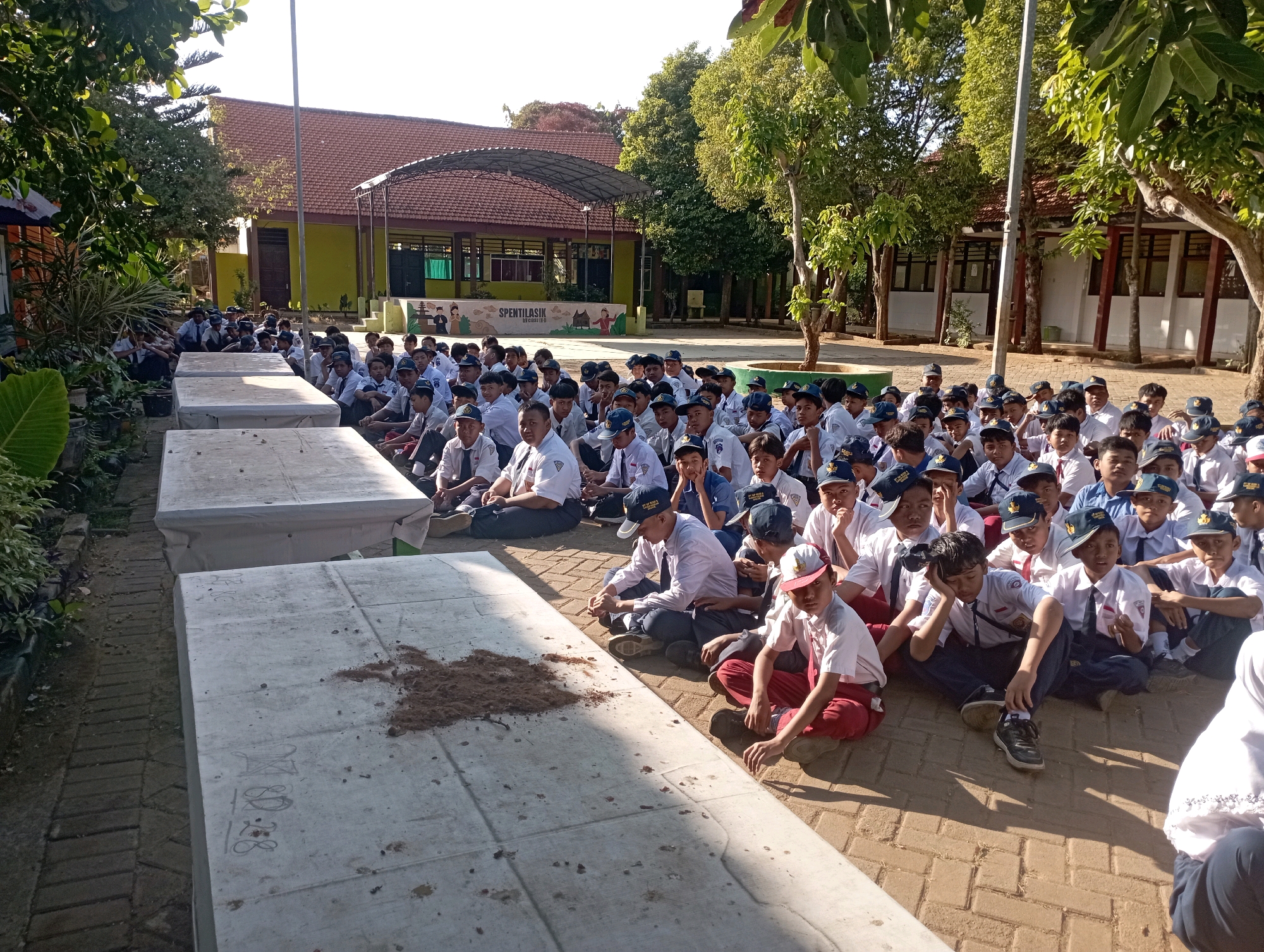The image depicts a large group of Indonesian school children, likely elementary or middle school-aged, sitting on a paved courtyard in what appears to be a school setting in Banyuurip, Indonesia. The children are dressed in their school uniforms, consisting of white shirts, dark pants or skirts, and blue or red hats. They are arranged in rows, facing towards the center of the courtyard. Some children are looking towards the camera, while others are engaged in conversation or have their heads down.
In the background, there is a school building with a red tiled roof and yellow walls, featuring covered walkways and windows. Trees are scattered around the courtyard, providing some shade. In the foreground, there are large, flat, concrete structures, possibly benches or platforms, some of which are covered with white tarpaulin. A pile of loose soil is visible on one of these structures. The ground is paved with reddish-brown bricks. The overall impression is of an outdoor school assembly or activity taking place on a sunny day.