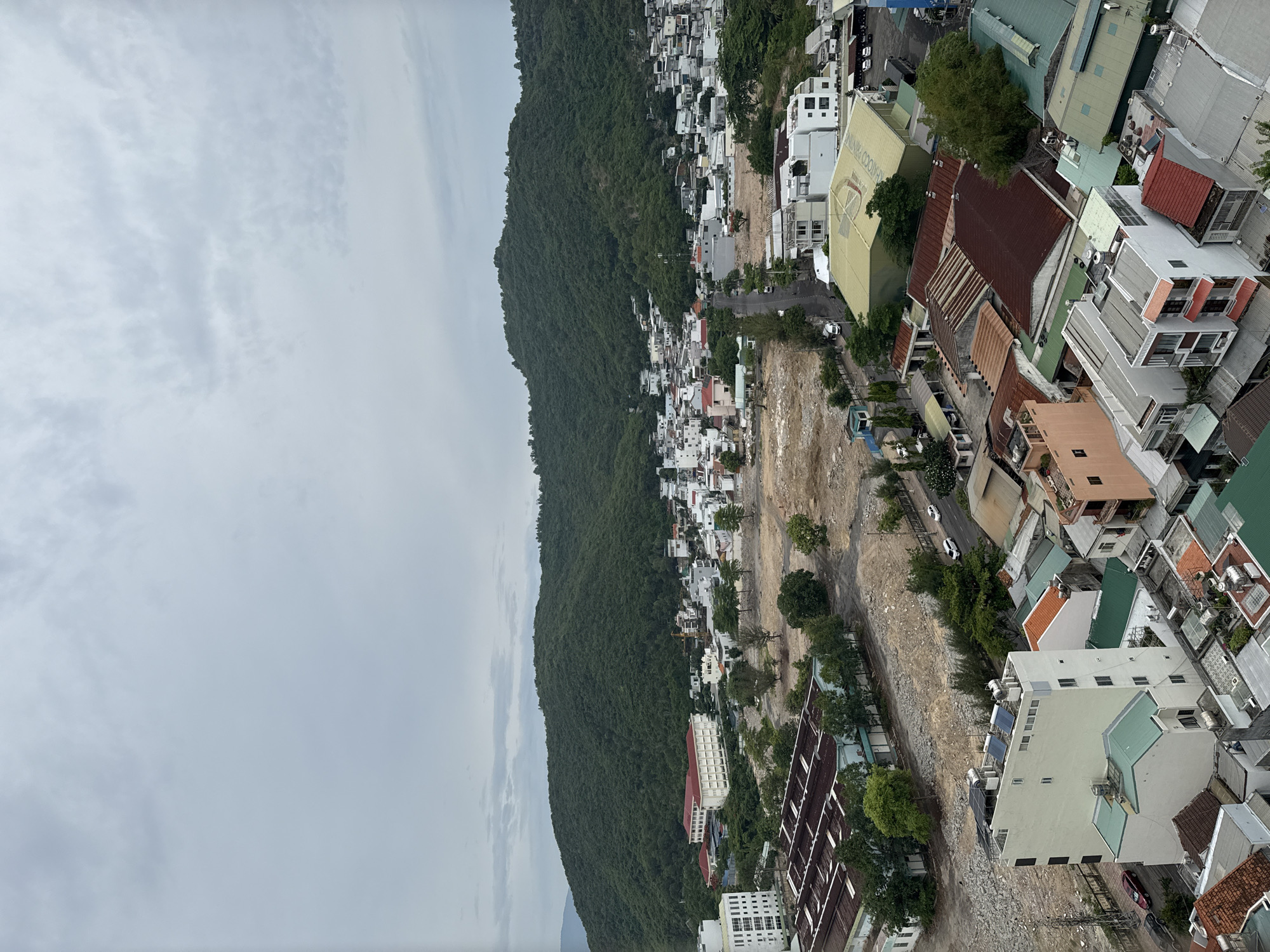 This is a high-angle aerial view of a city in Nha Trang, Vietnam, on a cloudy day. In the foreground, various buildings with different roof styles, including corrugated metal and tile, are visible. Some buildings are multi-story residential structures, while others appear to be commercial or industrial. A large, light-colored building with solar panels on its roof is prominently featured. Beyond these buildings, there is a large, undeveloped area of brown earth, possibly a construction site. Further in the distance, a dense, green, tree-covered mountain range dominates the background. A small cluster of buildings, with white and light-colored walls and red roofs, is nestled at the base of the mountain. A road with a few cars and a truck winds through the scene. The sky is overcast with a muted gray and white cloud cover.