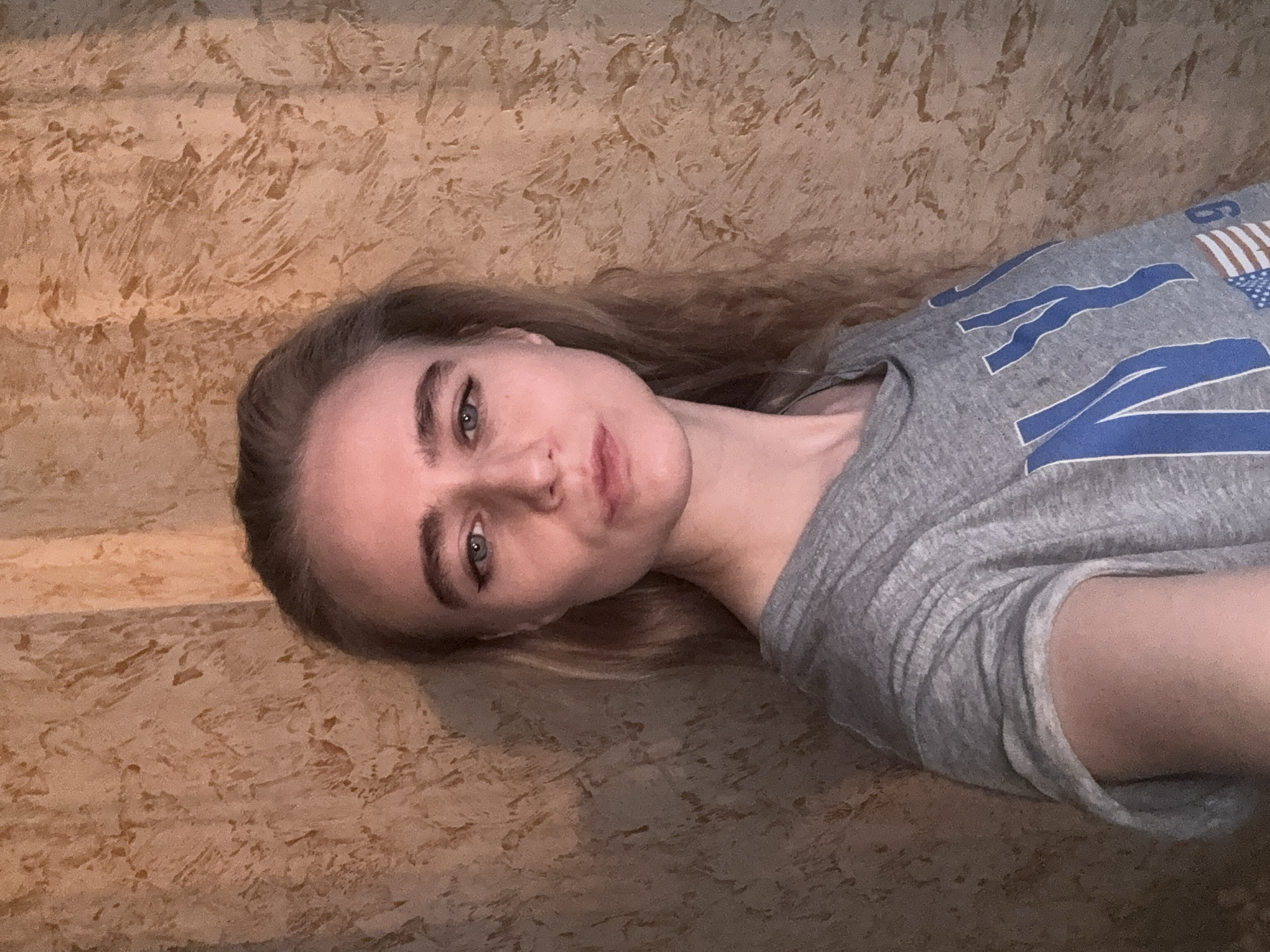 A young woman is depicted in a medium shot, lying down and looking directly at the camera. Her head is slightly tilted, and her long, light brown hair spreads out behind her. She has fair skin, distinct eyebrows, and green or blue eyes with visible eyeliner. Her expression is neutral and calm.
She is wearing a grey t-shirt that features a graphic design. Prominent blue letters, possibly spelling "NEW" or part of a larger word, are visible on the shirt. Next to the letters, there is a design resembling a partial American flag, with red and white stripes and a blue field.
The background is a textured wall or surface, characterized by a mottled pattern in varying shades of brown and tan, suggestive of stucco or a similar decorative finish. The lighting is soft and even, indicating an indoor setting. There are no clear cues for the time of day or specific location beyond being indoors.