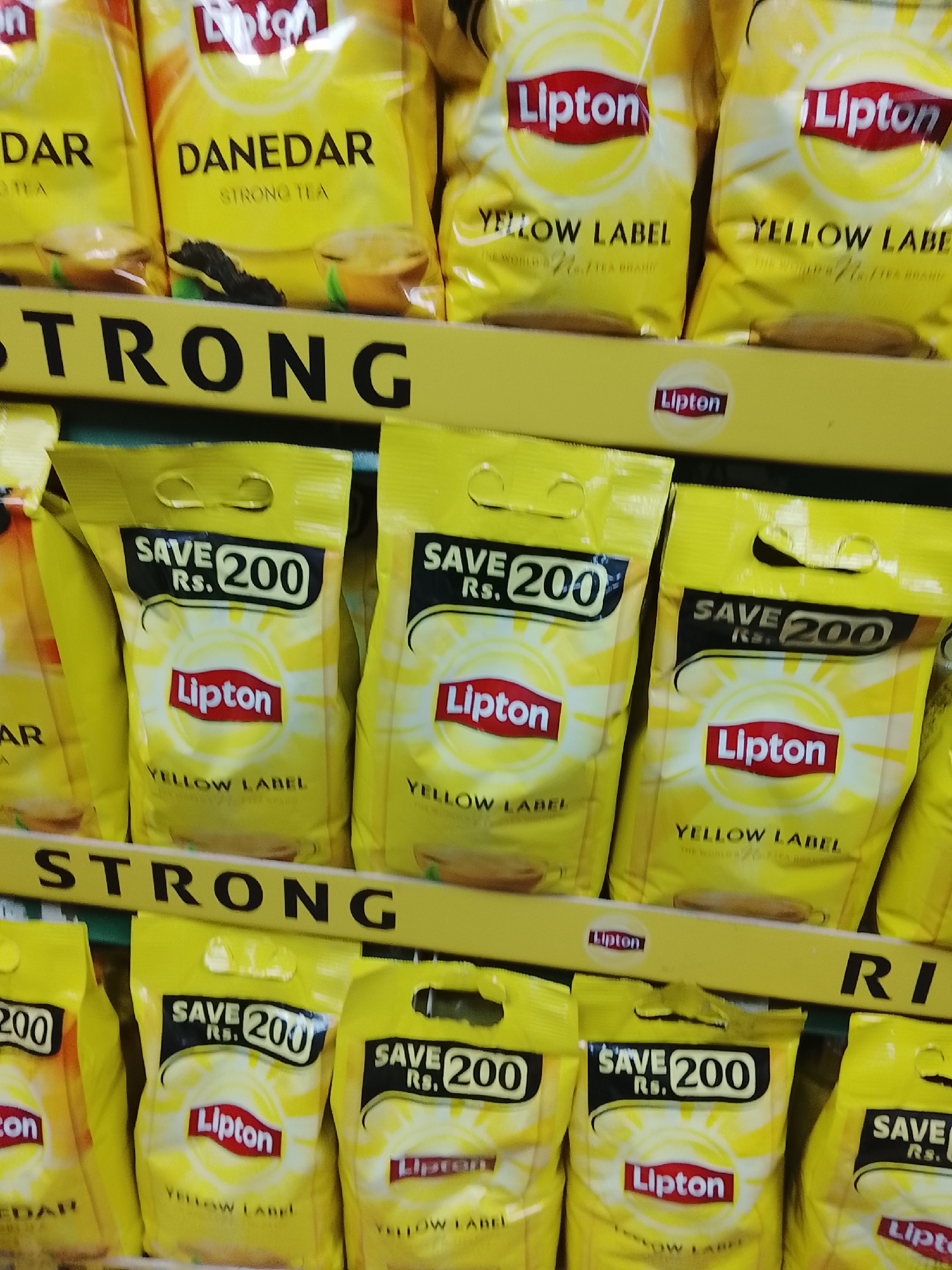 The image shows shelves stocked with Lipton tea packets in what appears to be a retail setting. The shelves are organized with yellow signage reading "STRONG" in black letters. There are two types of Lipton tea visible: "Danedar Strong Tea" and "Yellow Label". The Yellow Label tea packets feature prominent "SAVE Rs. 200" messaging. The location is not explicitly mentioned in the image, but it is implied in the prompt that it may be Gulgasht Colony, Pakistan.