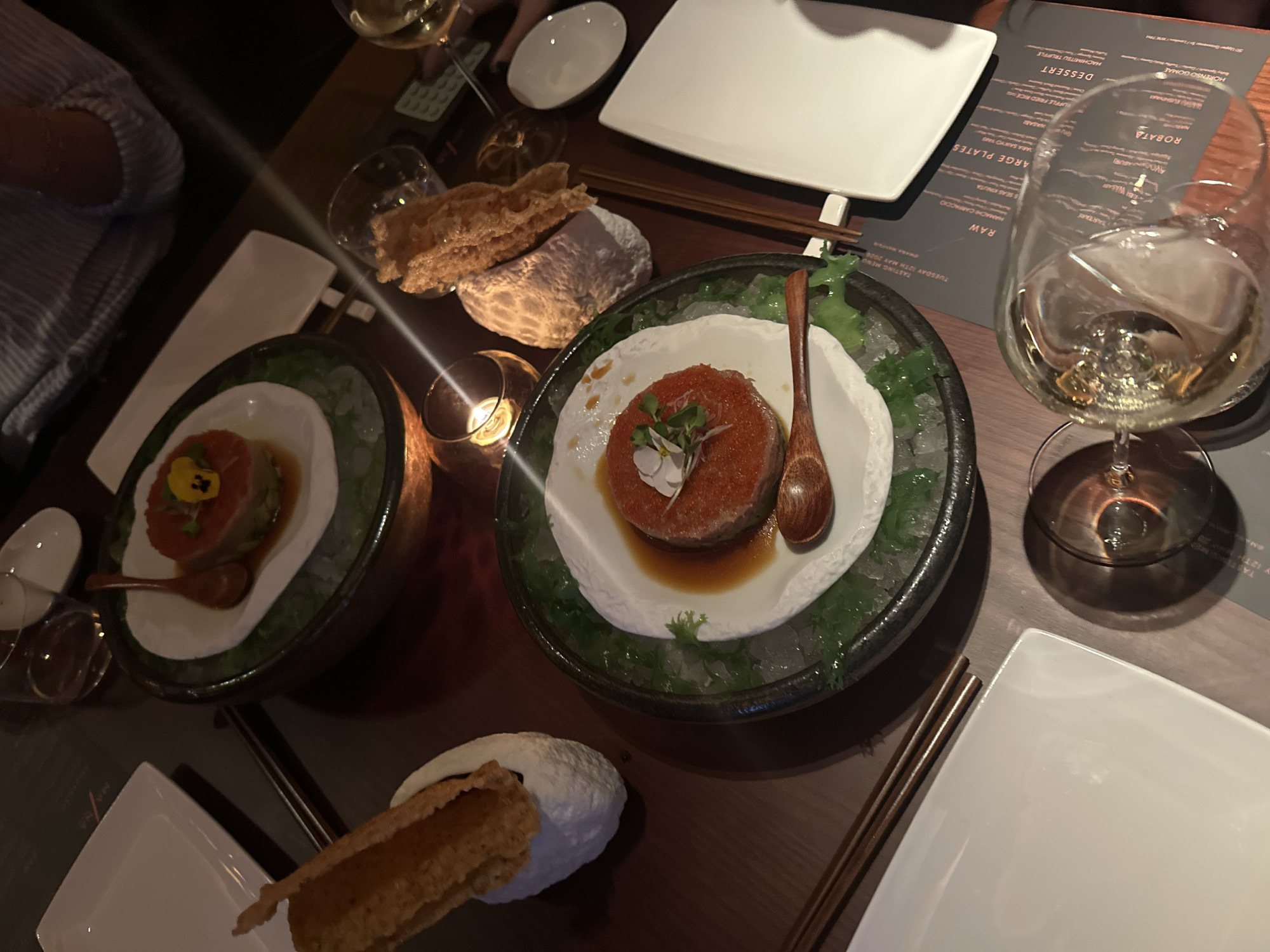Here is a detailed description of the image:
The image shows a dining table at what appears to be a high-end restaurant, judging by the presentation and quality of the food. The scene is somewhat dark, with focused lighting on the dishes.
Several plates of food are visible. There are two round plates, each containing a mound of what seems to be raw fish or tartare on a bed of ice and garnished with greens and a small flower. There are also crispy-looking fried elements accompanied by white creamy mounds, possibly dips or sauces.
Alongside the food are square white plates, chopsticks, and a menu card. The menu's text includes the words "TASTING MENU", "TARTARE", "RAW", "ROBATA", "DESSERT," along with Japanese dishes like "HORENSO GOMAE".
Wine glasses, one partially full with a light-colored liquid, and a small candle in a votive holder can also be seen. A person’s arm and part of their patterned shirt is visible in the upper left corner.
The restaurant is likely located in London, United Kingdom. The menu indicates "TUESDAY 12TH MAY 2006" and notes at the very top edge suggest the restaurant name and address are "...TAY BY 127 L 29 LONDON..."
